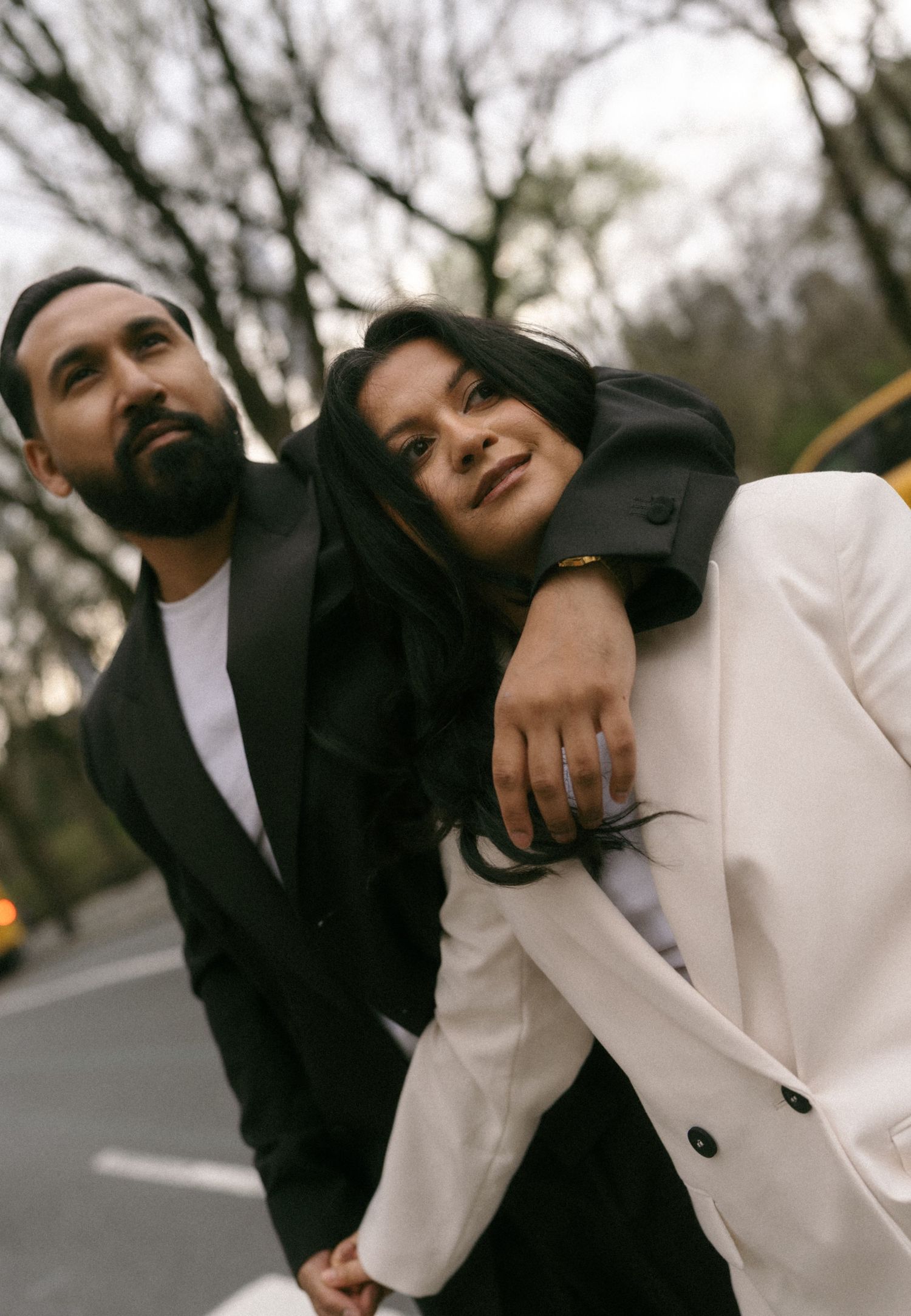 Stylish couple in black and white blazers posing outdoors in a park setting with bare trees in the background.
