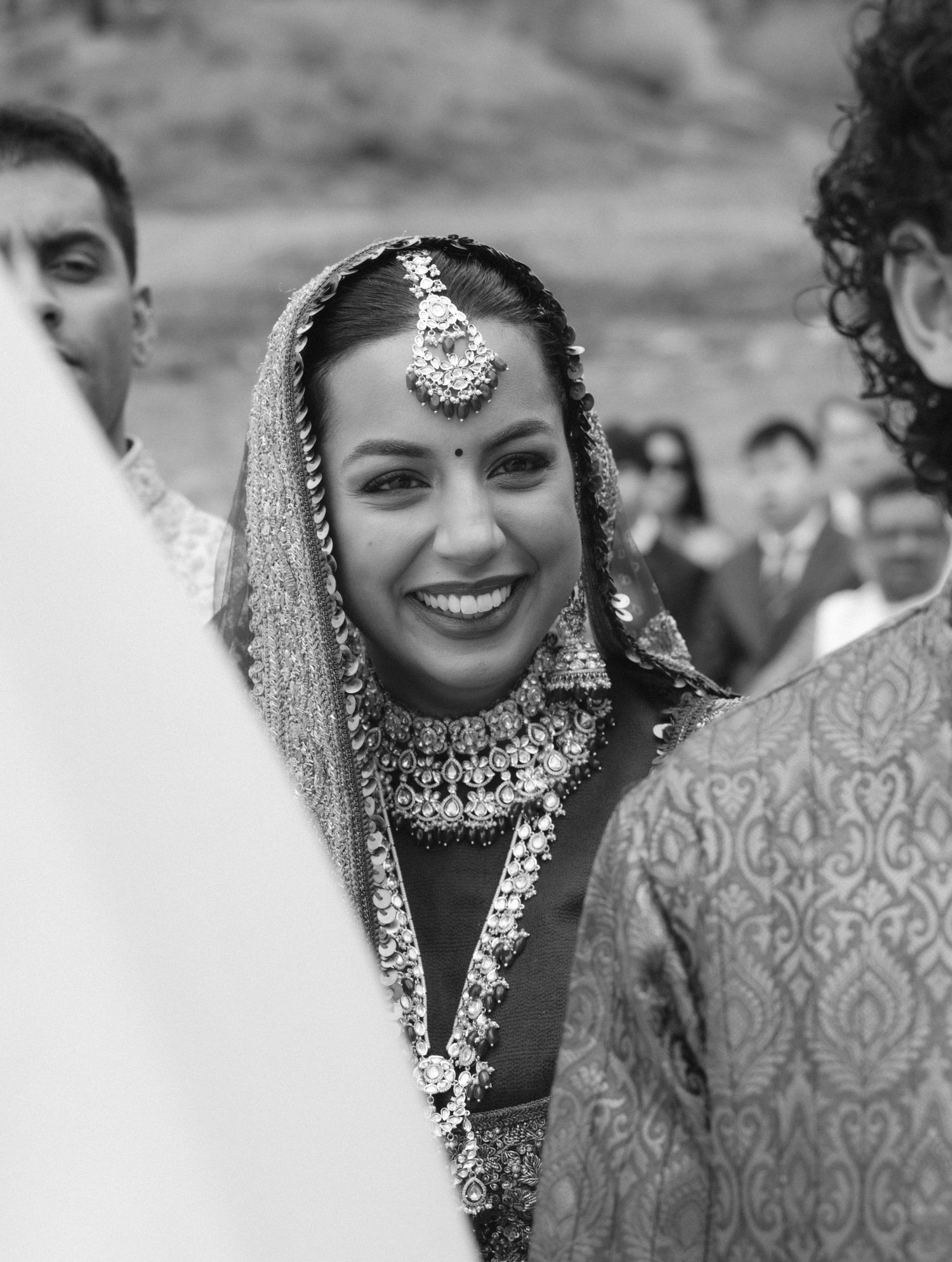 Smiling Indian bride in traditional jewelry and dupatta during wedding ceremony, black and white photo.