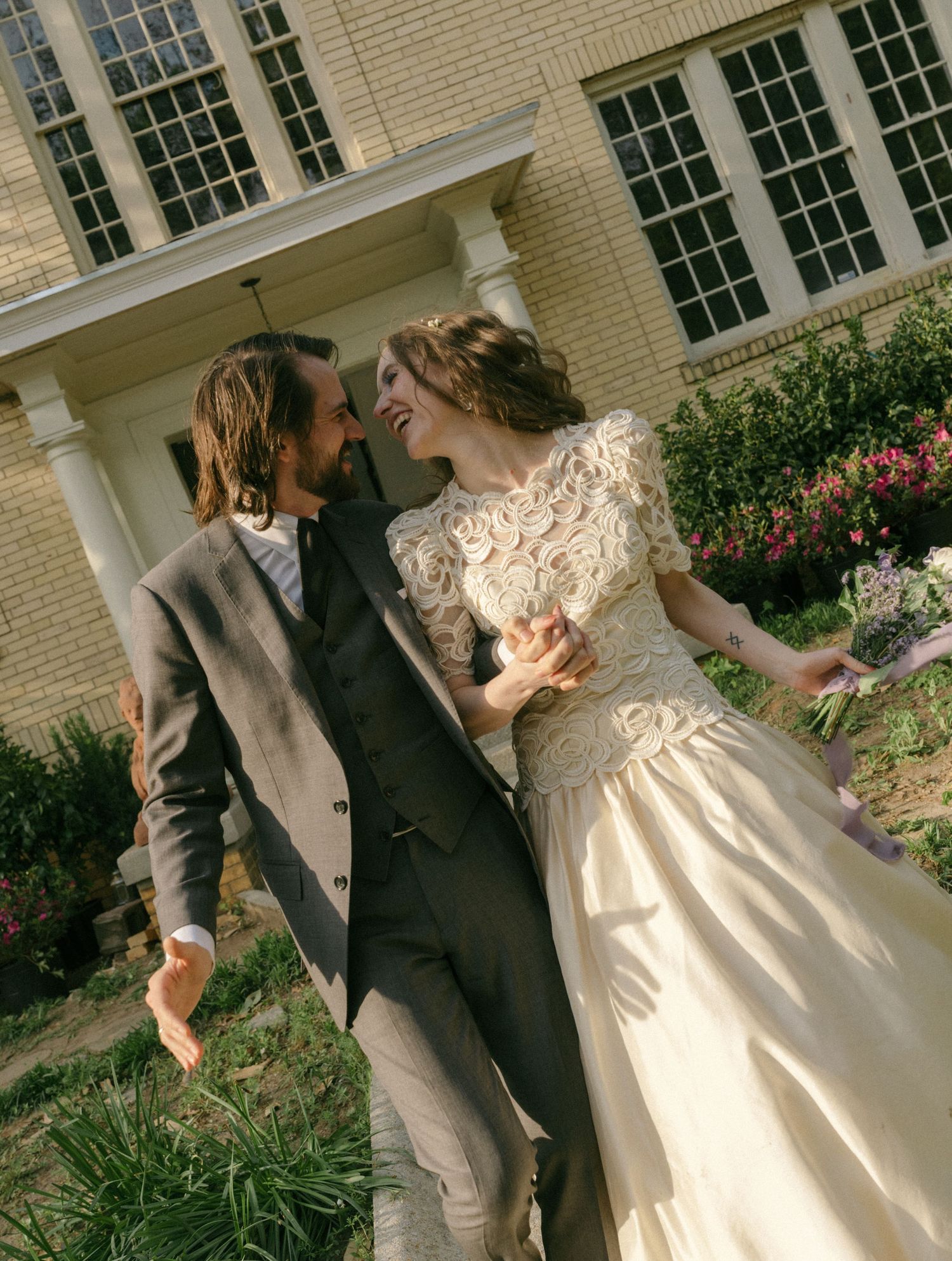 A joyful bride in a vintage lace gown and groom in a gray suit walk together outside a classic brick building.