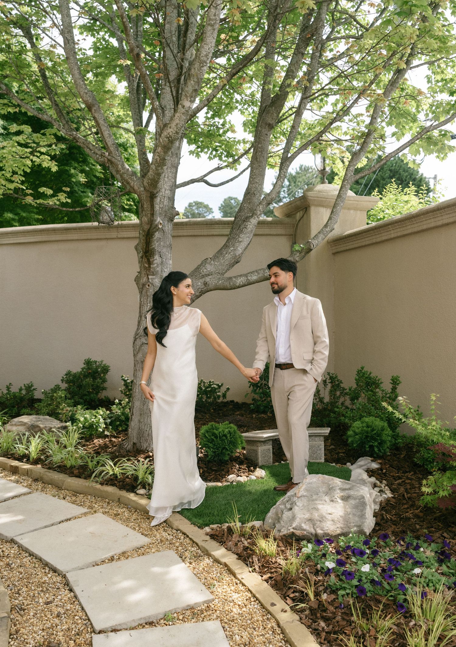 Couple in wedding attire holding hands in a lush garden with stone path, large tree, and flowering plants.