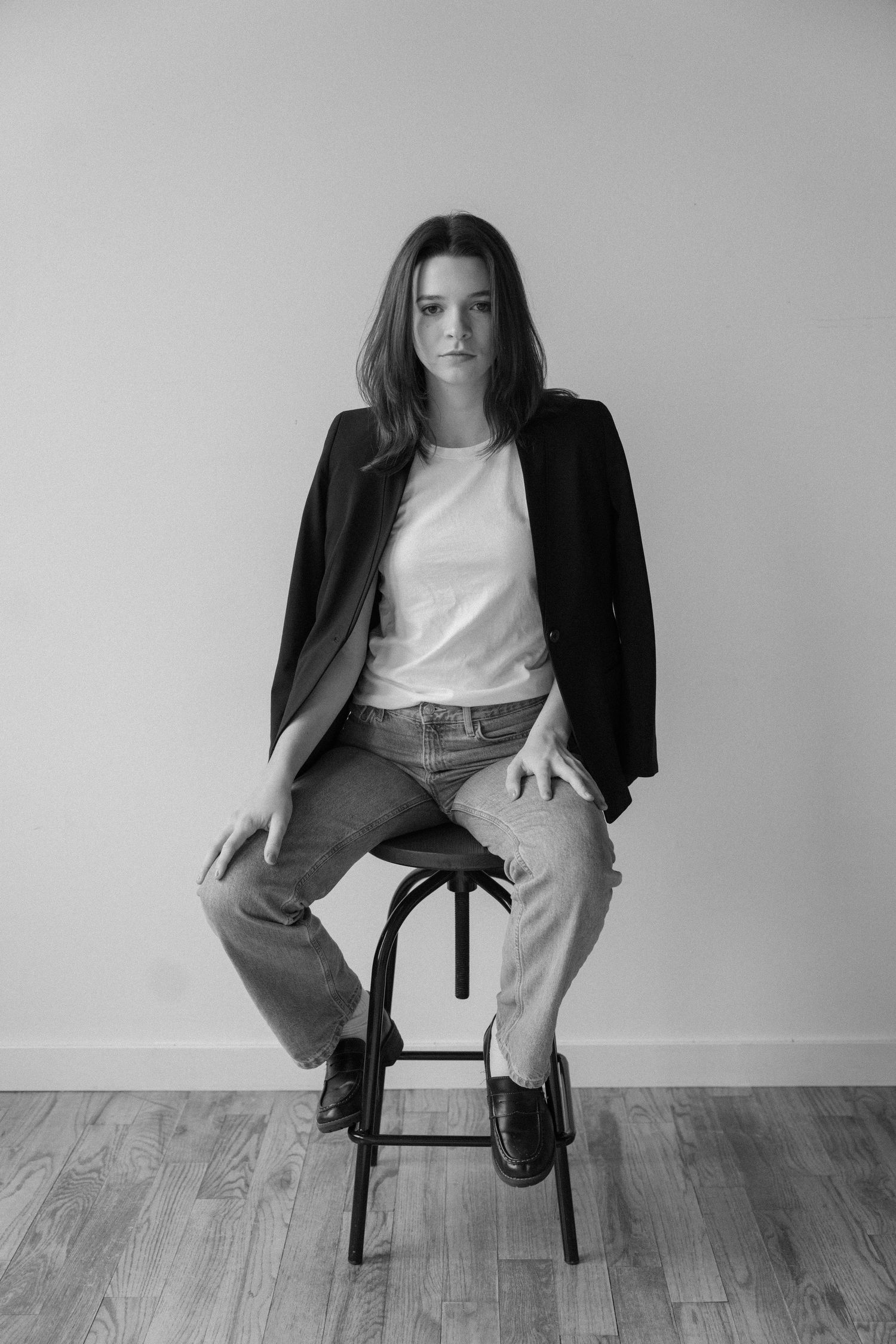 Black and white portrait of a young woman in blazer and jeans sitting on a stool against a plain wall.