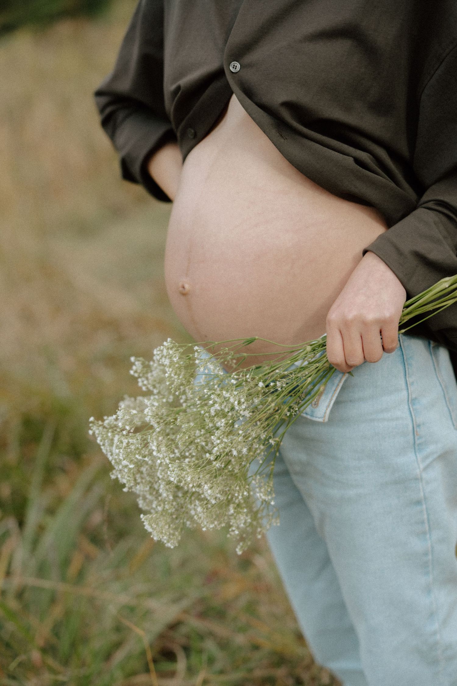 Pregnant woman in dark shirt and jeans holding white wildflowers bouquet outdoors in a field.