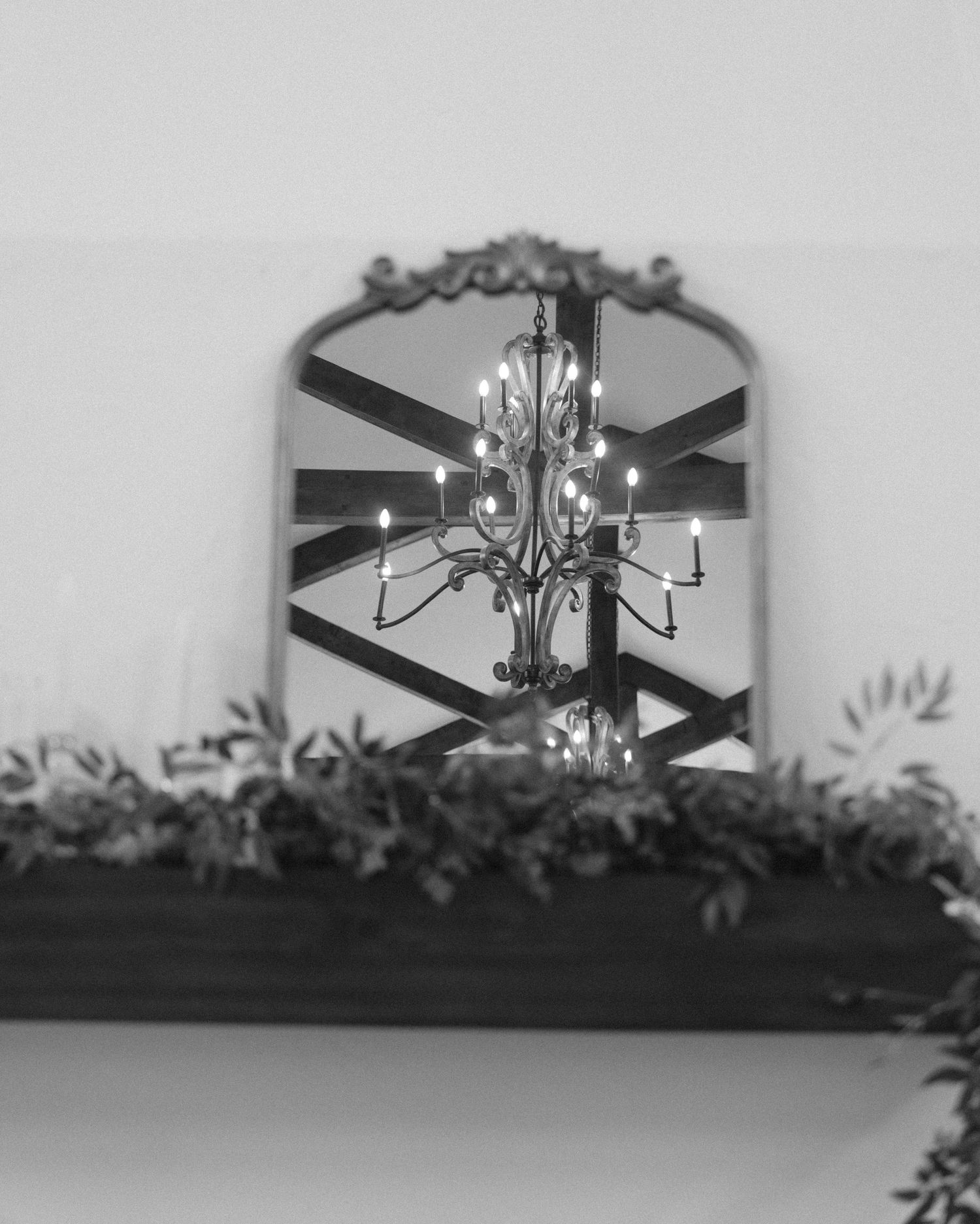 Ornate vintage mirror reflecting an elegant chandelier above a mantle decorated with greenery.