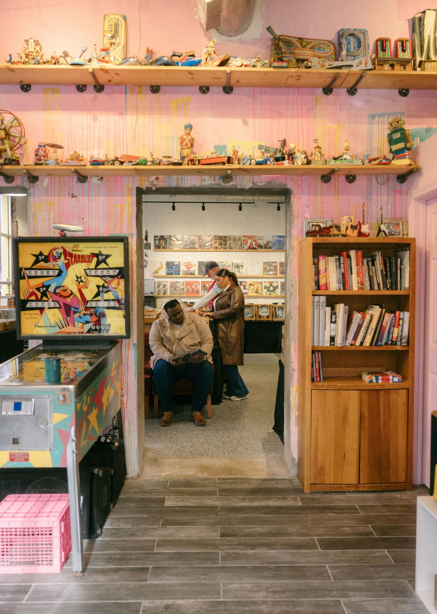 Colorful retro shop with pinball machine, toy-lined shelves, bookcase, and two people visible through doorway in back room.