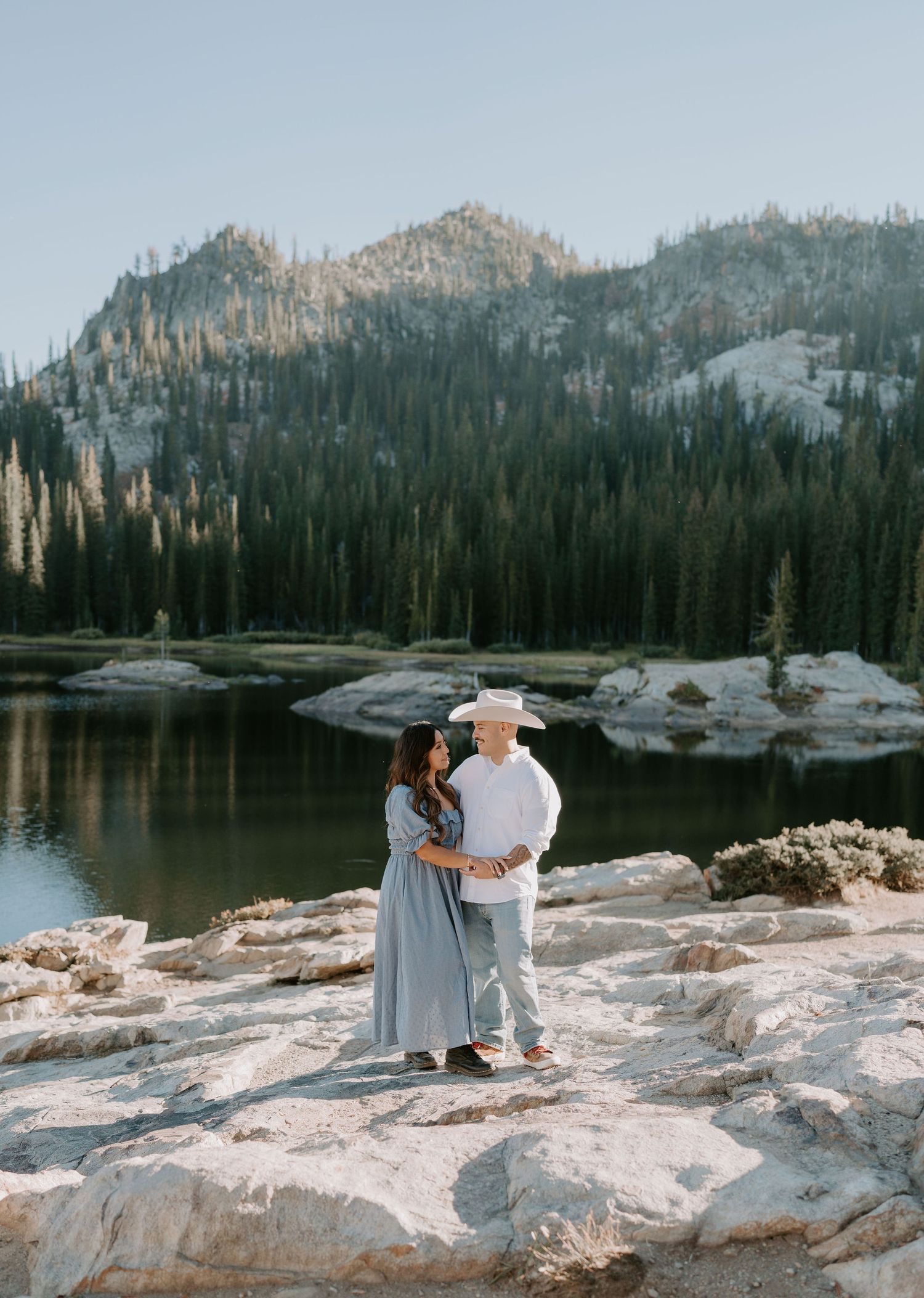 Couple embracing on rocky shore of mountain lake surrounded by pine trees and peaks in a scenic outdoor engagement photo.