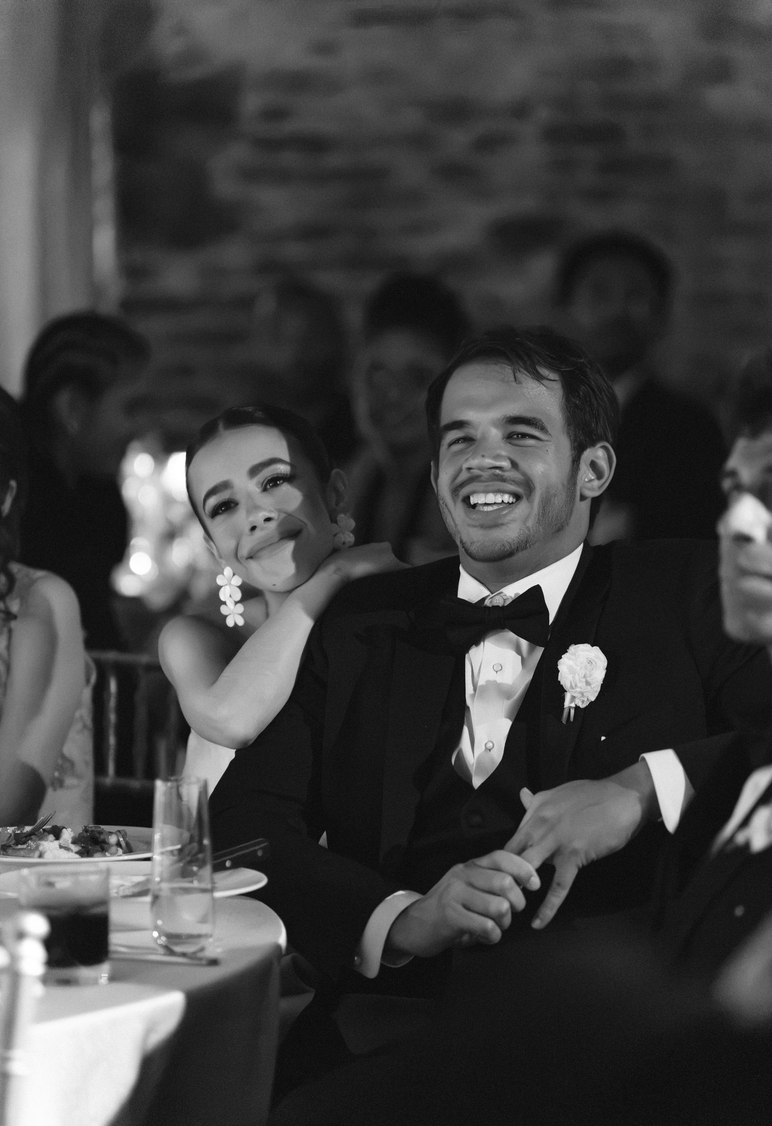 Black and white photo of a smiling groom in a tuxedo sitting beside a laughing bride at their wedding reception.