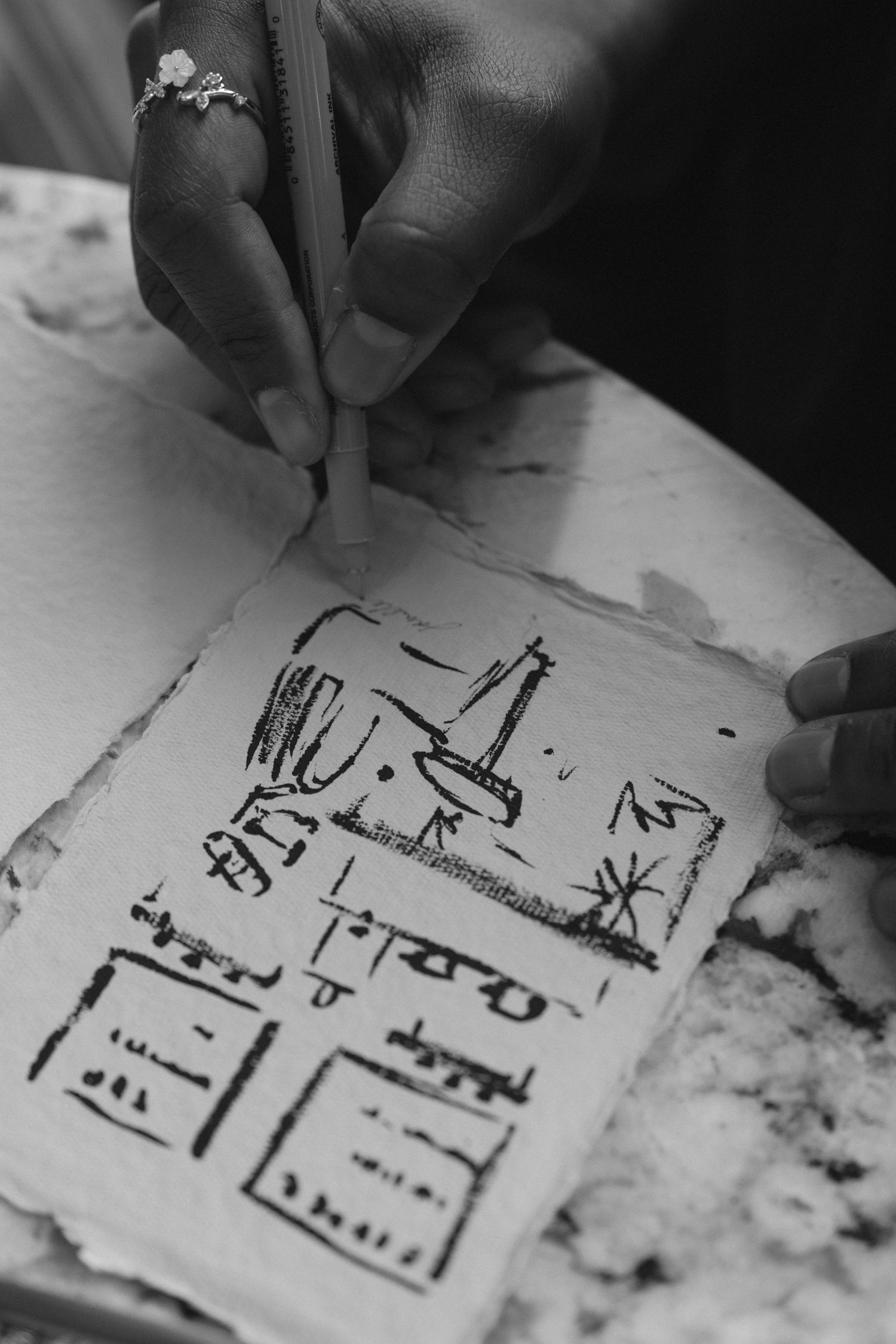 A dark-skinned hand with a ring sketches symbols on textured paper with a pencil in black and white.