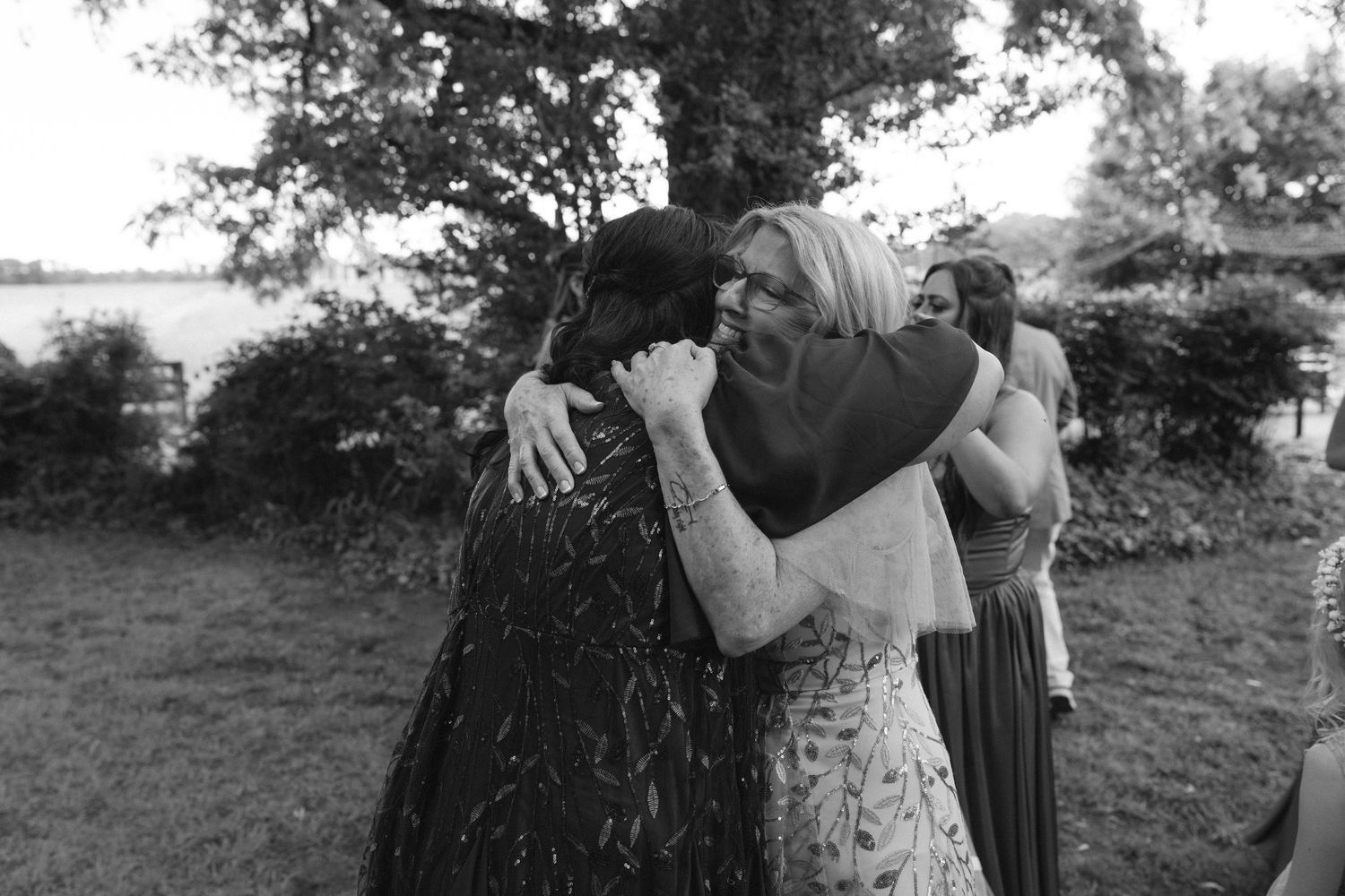 Black and white photo of guests sharing emotional hugs at an outdoor wedding ceremony near a lake.