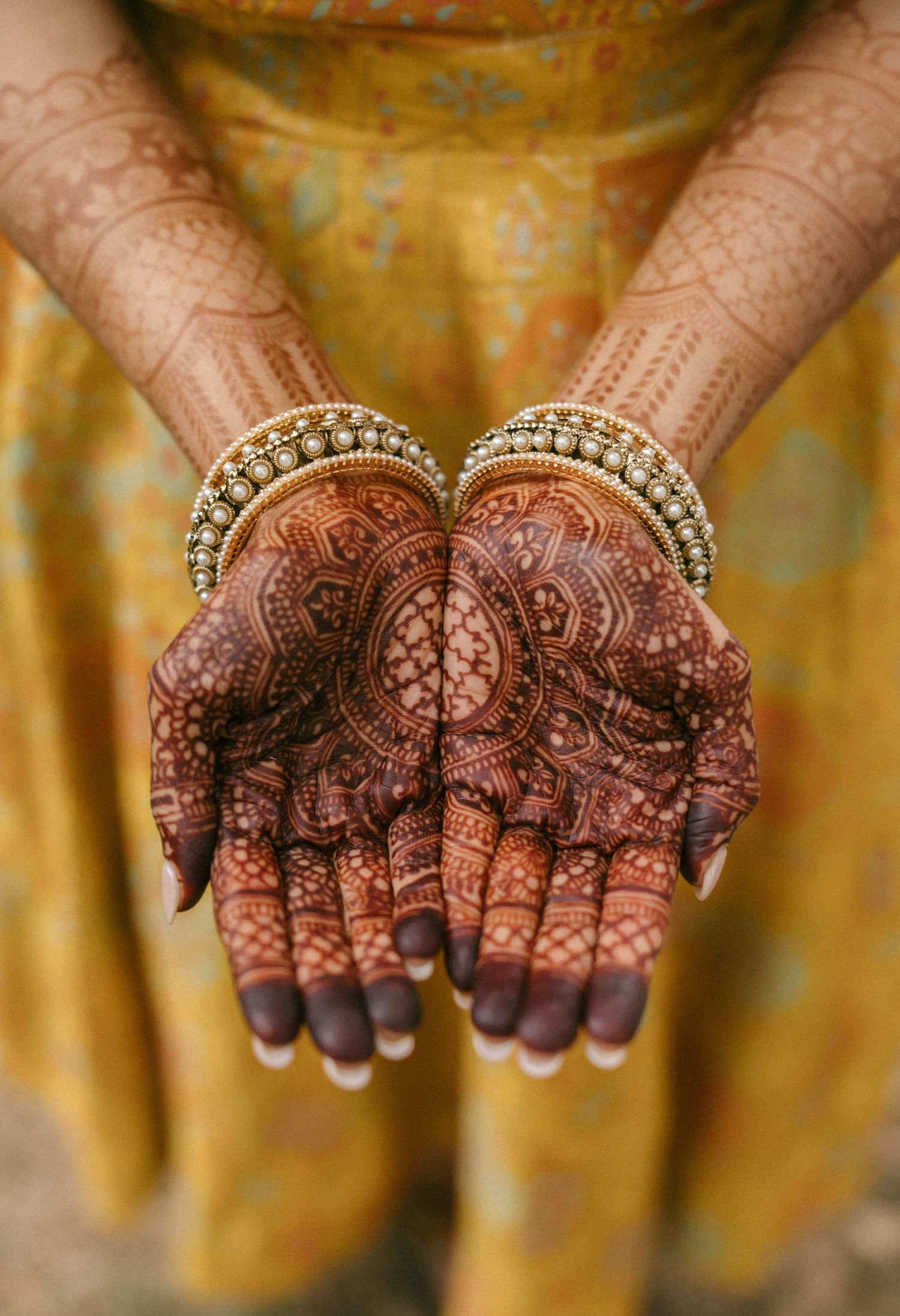 Close-up of Indian bride's palms displaying detailed mehndi henna patterns with gold bangles against yellow outfit.