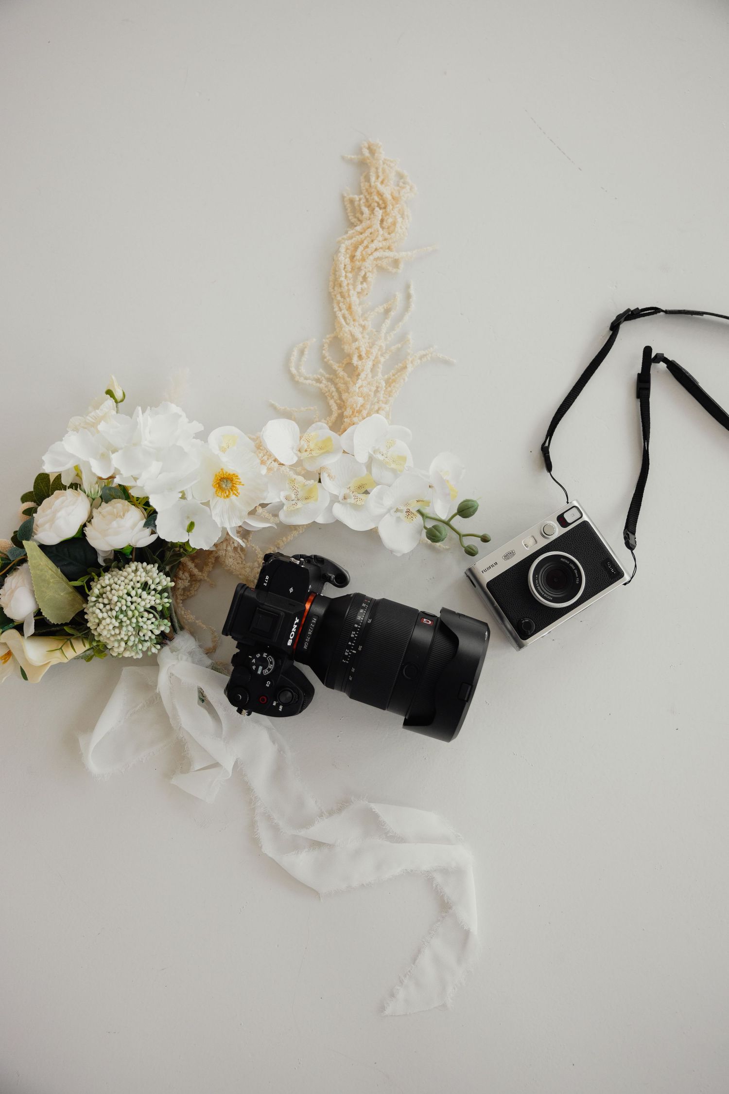 Flat lay of two cameras, white orchid bouquet, and a decorative feather arrangement on a light gray background.