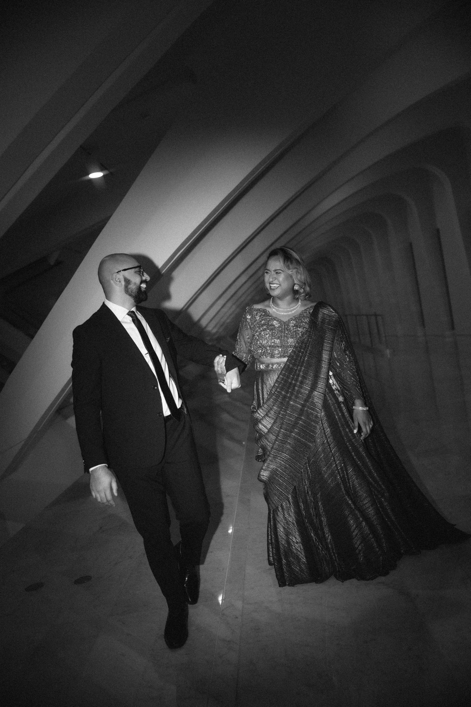 Black and white photo of an elegant couple walking together, man in suit and woman in traditional Indian saree.