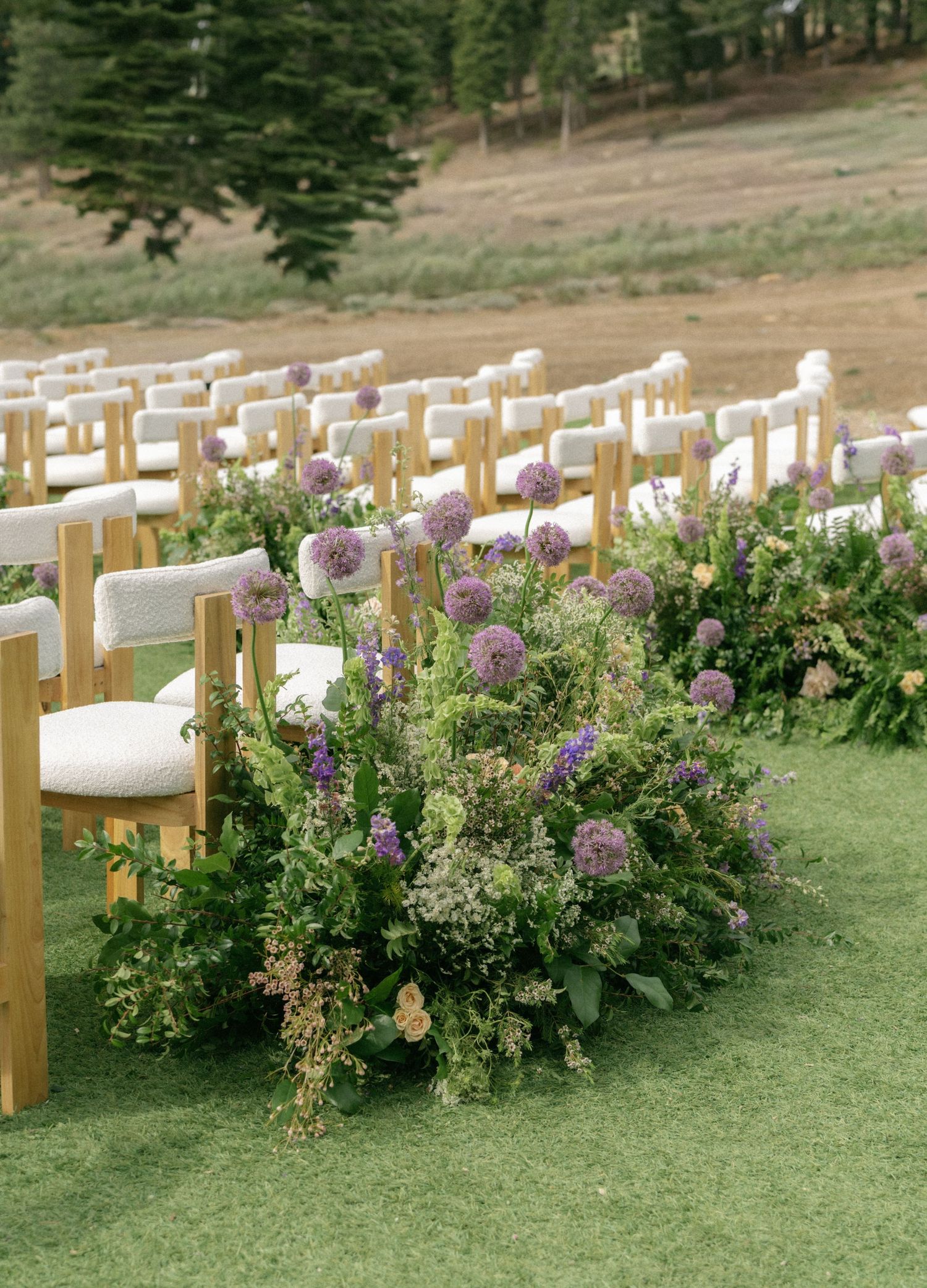 Outdoor wedding ceremony chairs lined with lush purple allium and wildflower arrangements on green grass aisle.
