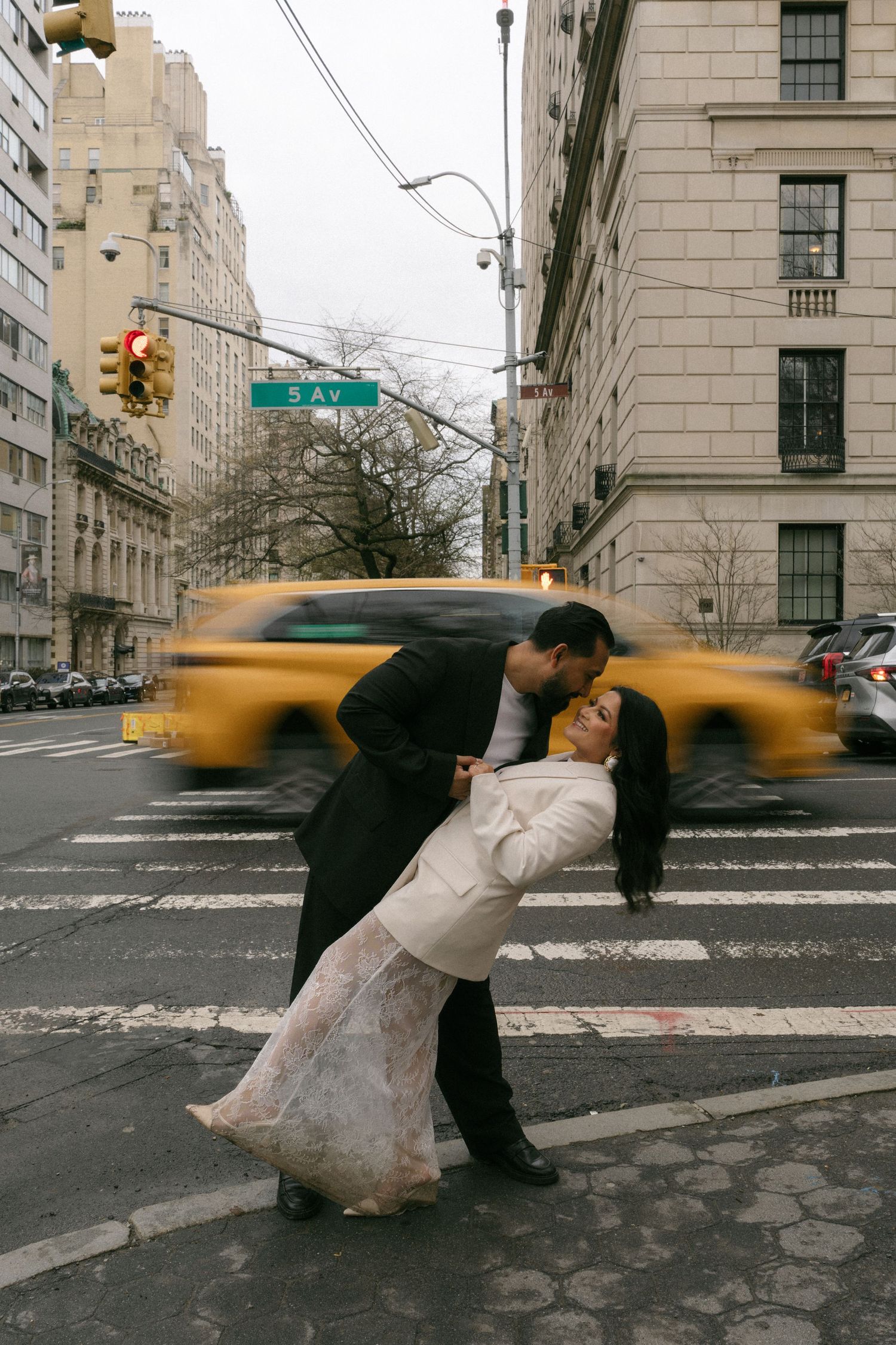Couple sharing a romantic dip kiss on 5th Avenue crosswalk with yellow NYC taxi passing.