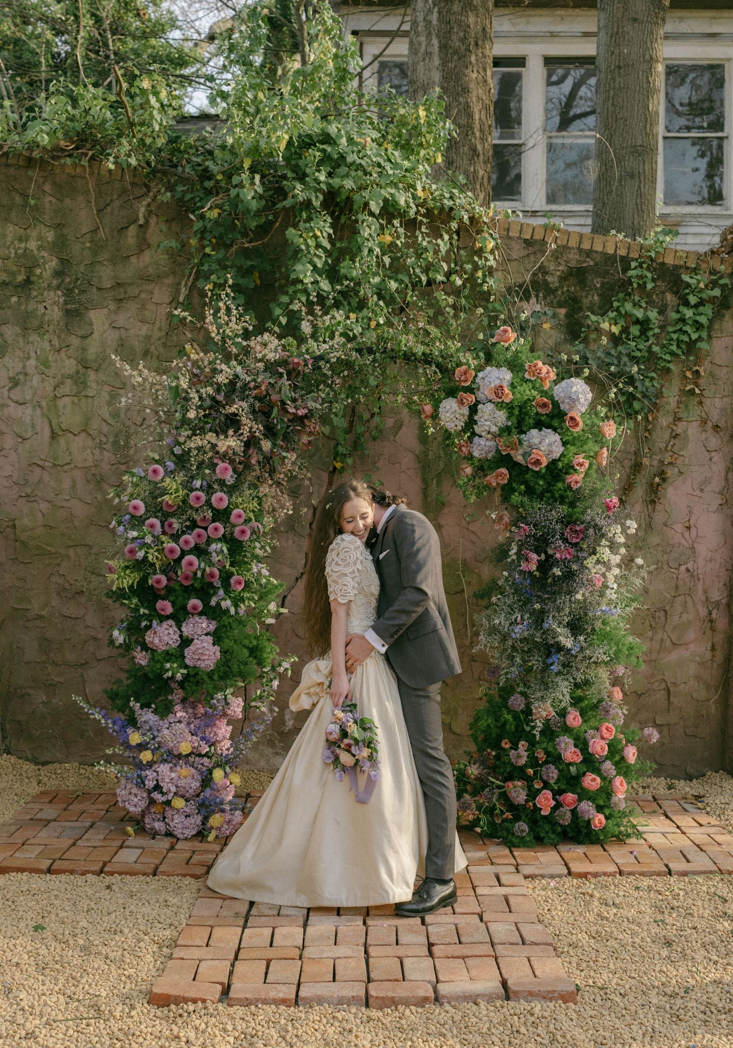 Bride and groom sharing a kiss under a lush floral arch with pink and purple blooms at an outdoor wedding ceremony.