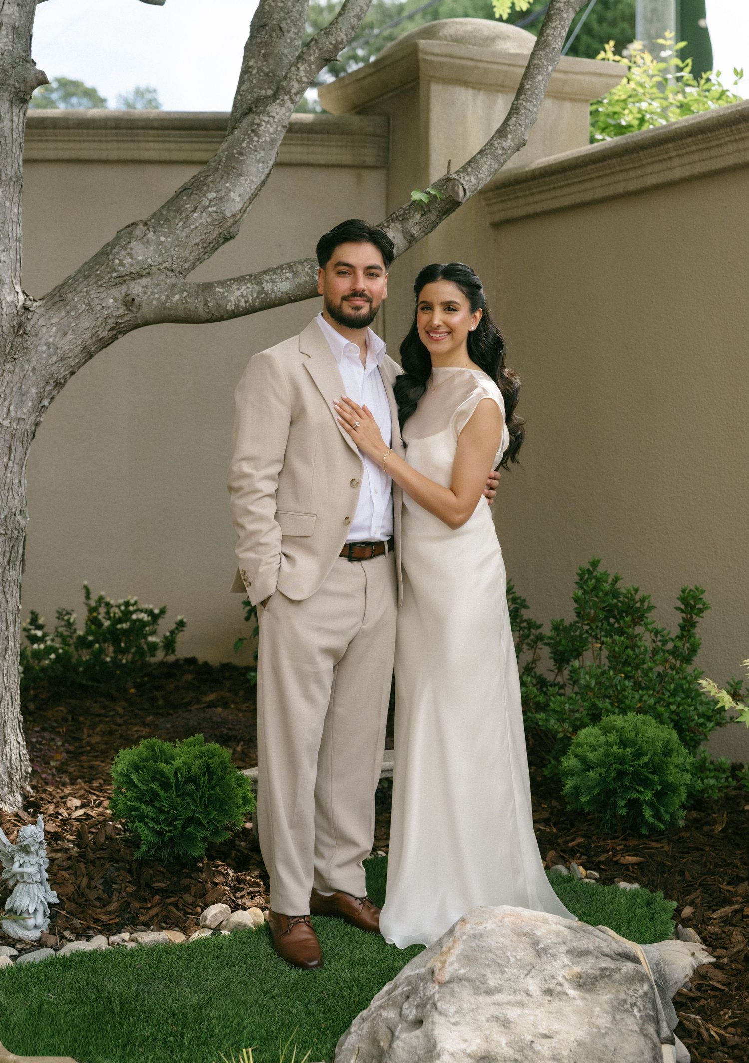 Couple in wedding attire posing outdoors, groom in beige suit and bride in white gown, standing near a tree.