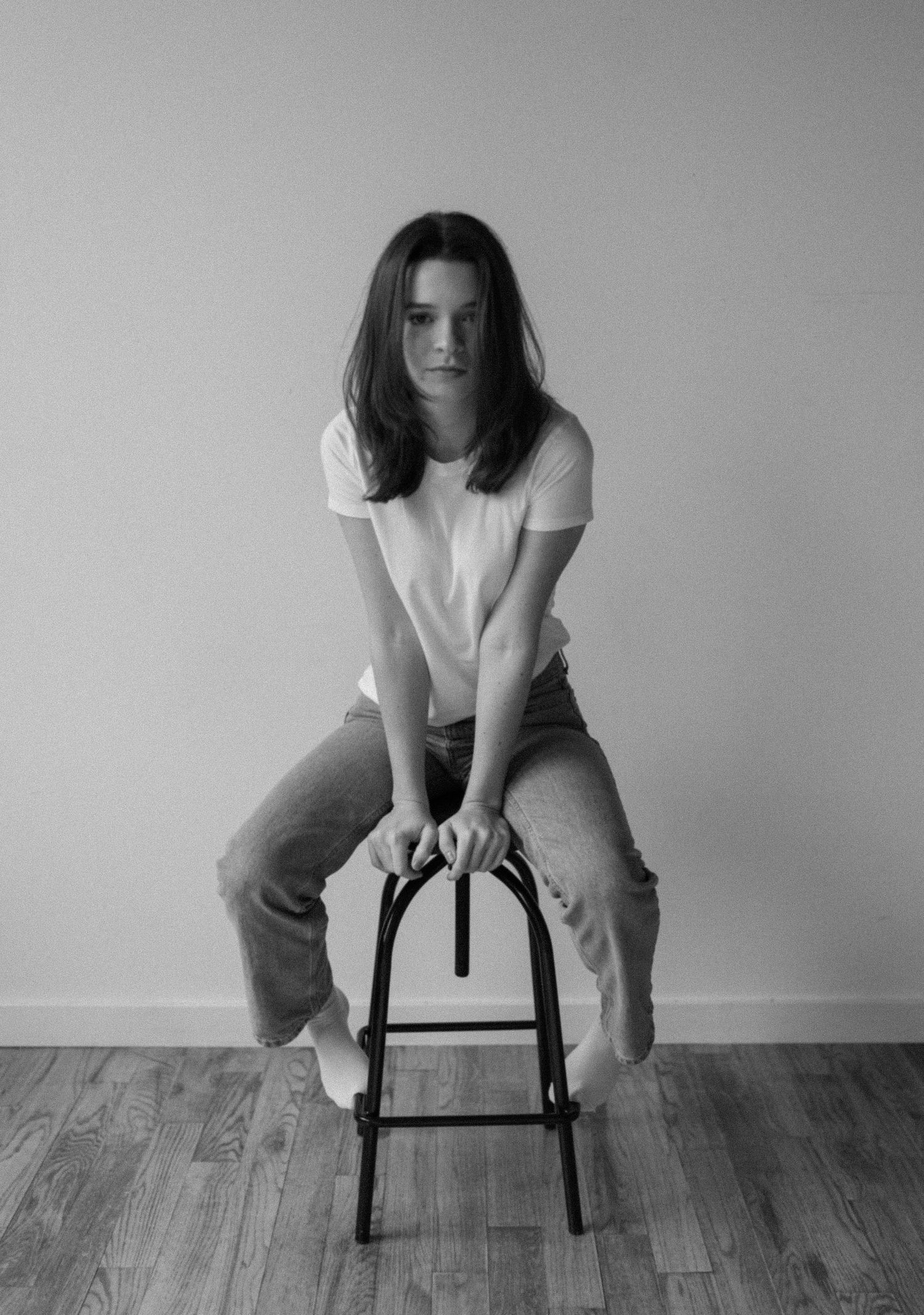 Black and white photo of a young woman in white tee and jeans, leaning on a stool on hardwood floor.