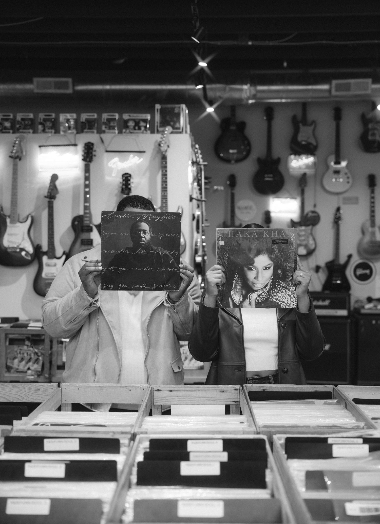 Two people browse vinyl records in a music shop surrounded by electric guitars mounted on the wall.