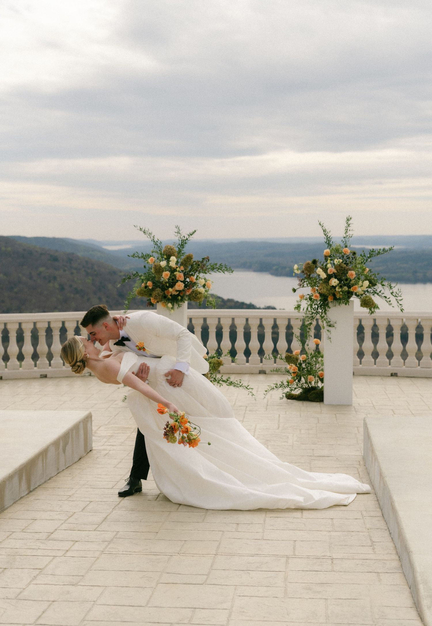 Groom dips bride for a romantic kiss on a stone terrace overlooking a river valley, framed by floral arrangements.