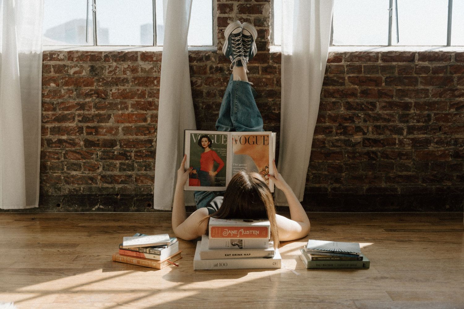 Woman lying on hardwood floor surrounded by stacked books and magazines, reading against a sunlit brick wall.