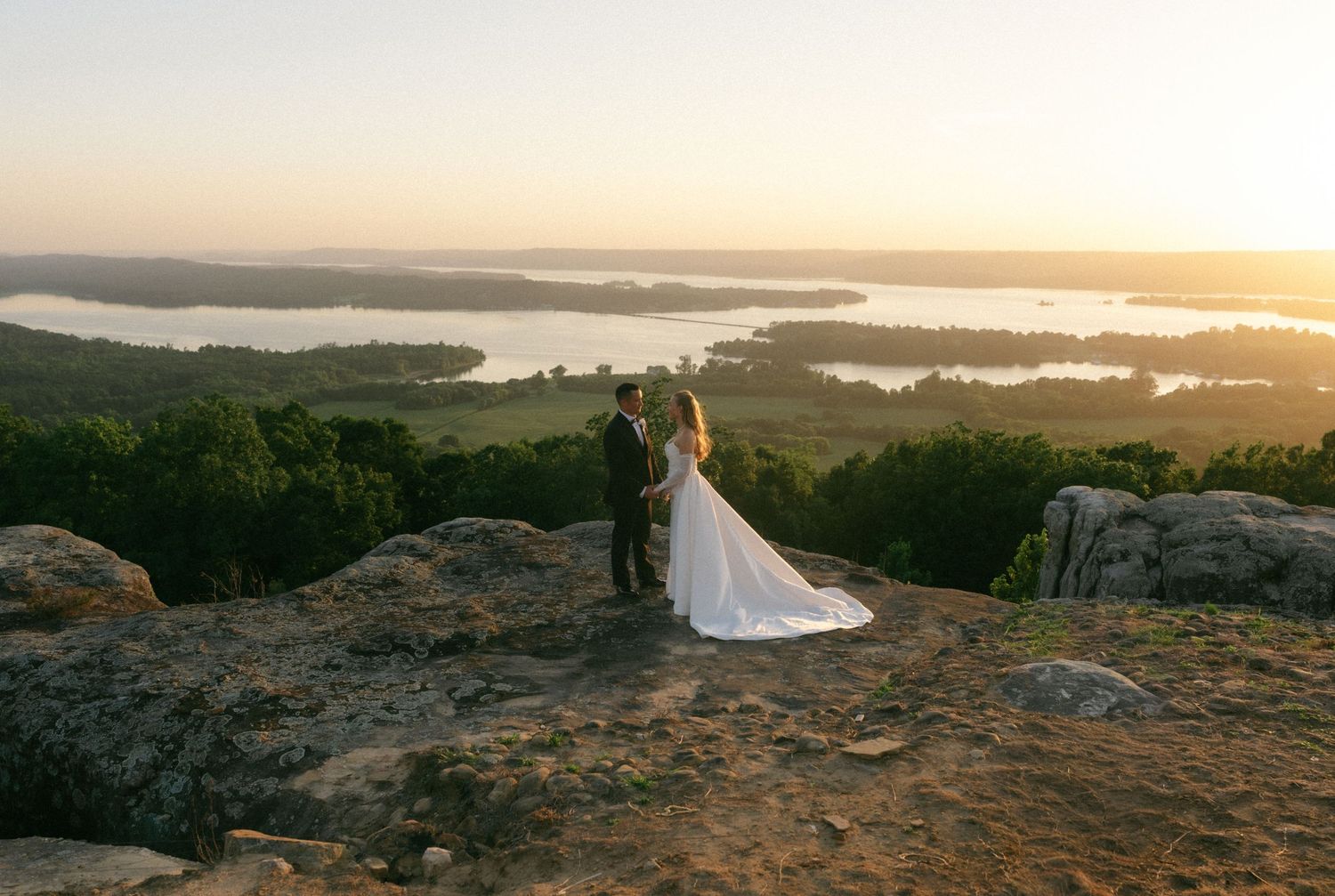 Bride and groom embrace on rocky hilltop at sunset with panoramic lake and forest views below.