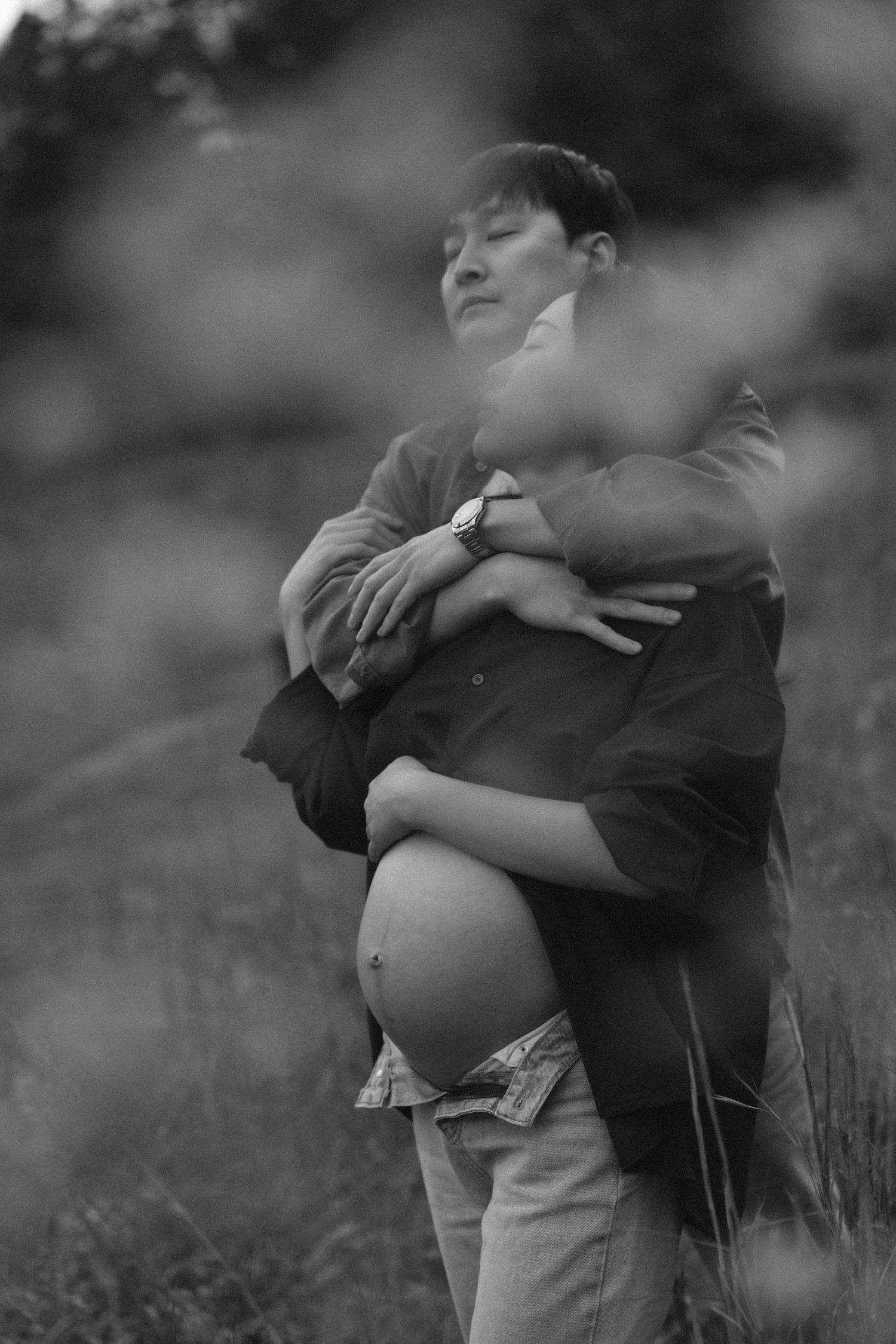 Black and white photo of a man embracing a pregnant woman from behind in a field, both eyes closed peacefully.