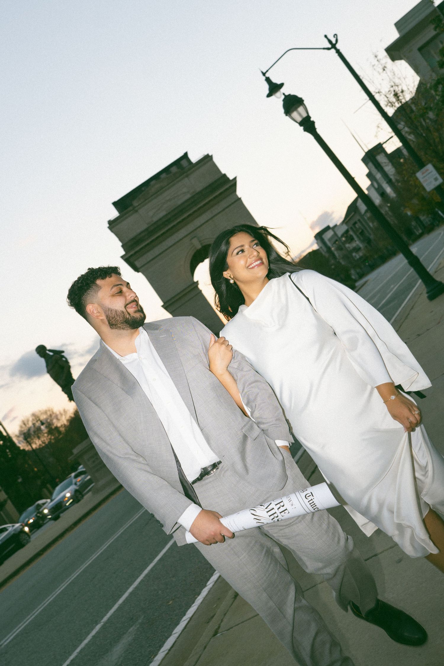 A smiling couple in elegant attire walks near a triumphal arch at dusk, man in gray suit, woman in white dress.
