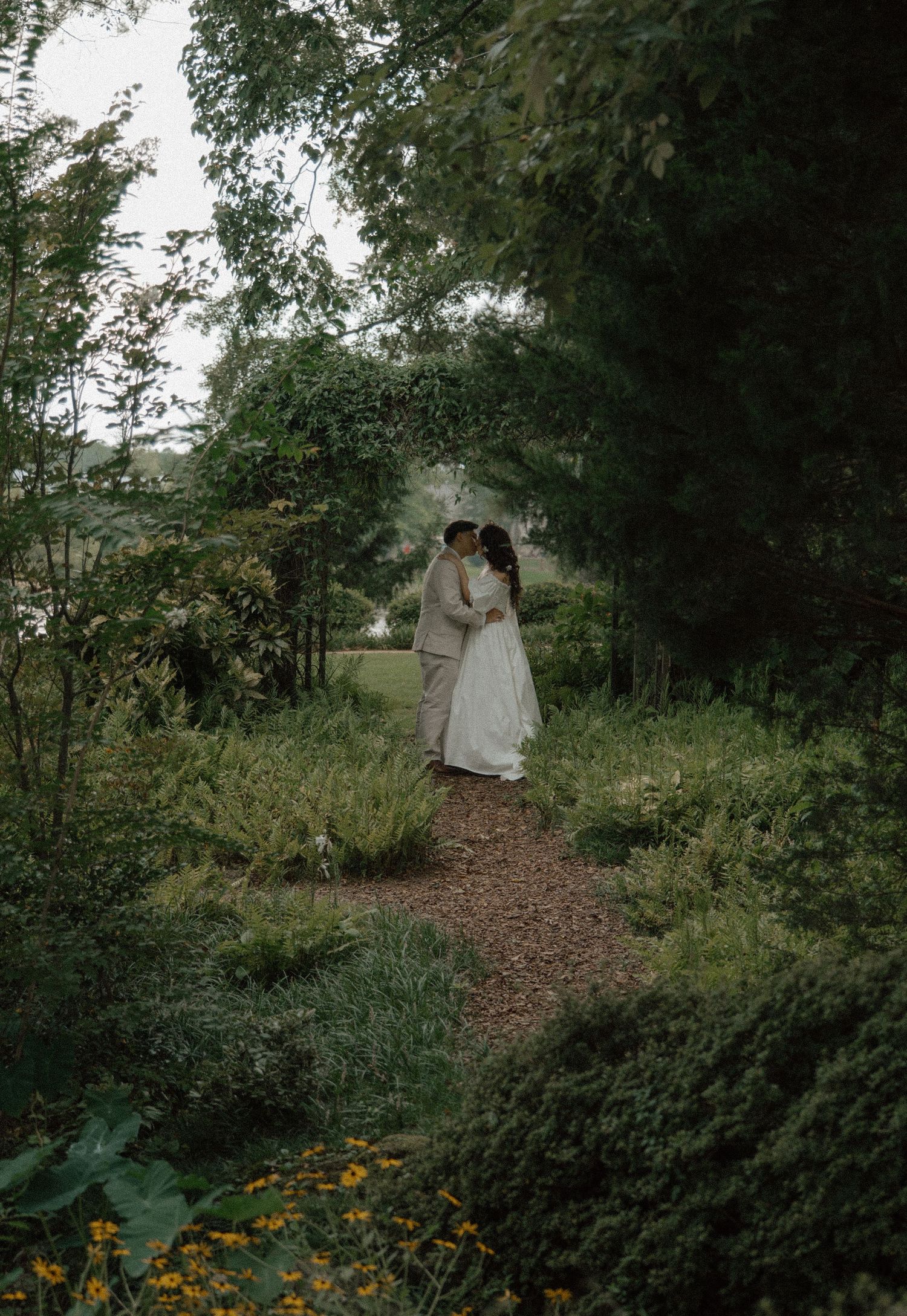 A couple shares a romantic kiss on a garden path surrounded by lush greenery and yellow wildflowers on their wedding day.