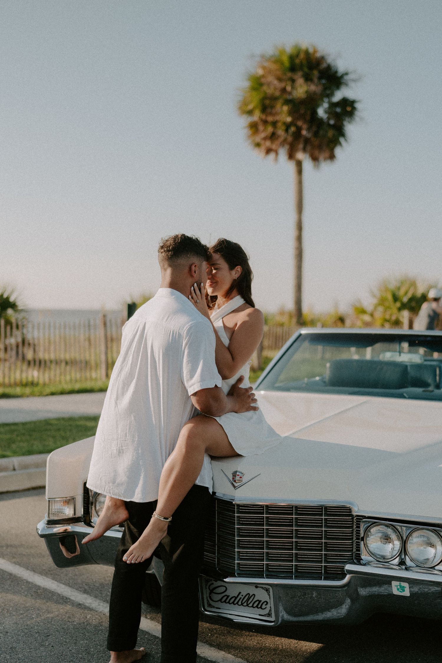 Couple sharing a romantic moment on a vintage white Cadillac convertible near a palm tree and ocean view.