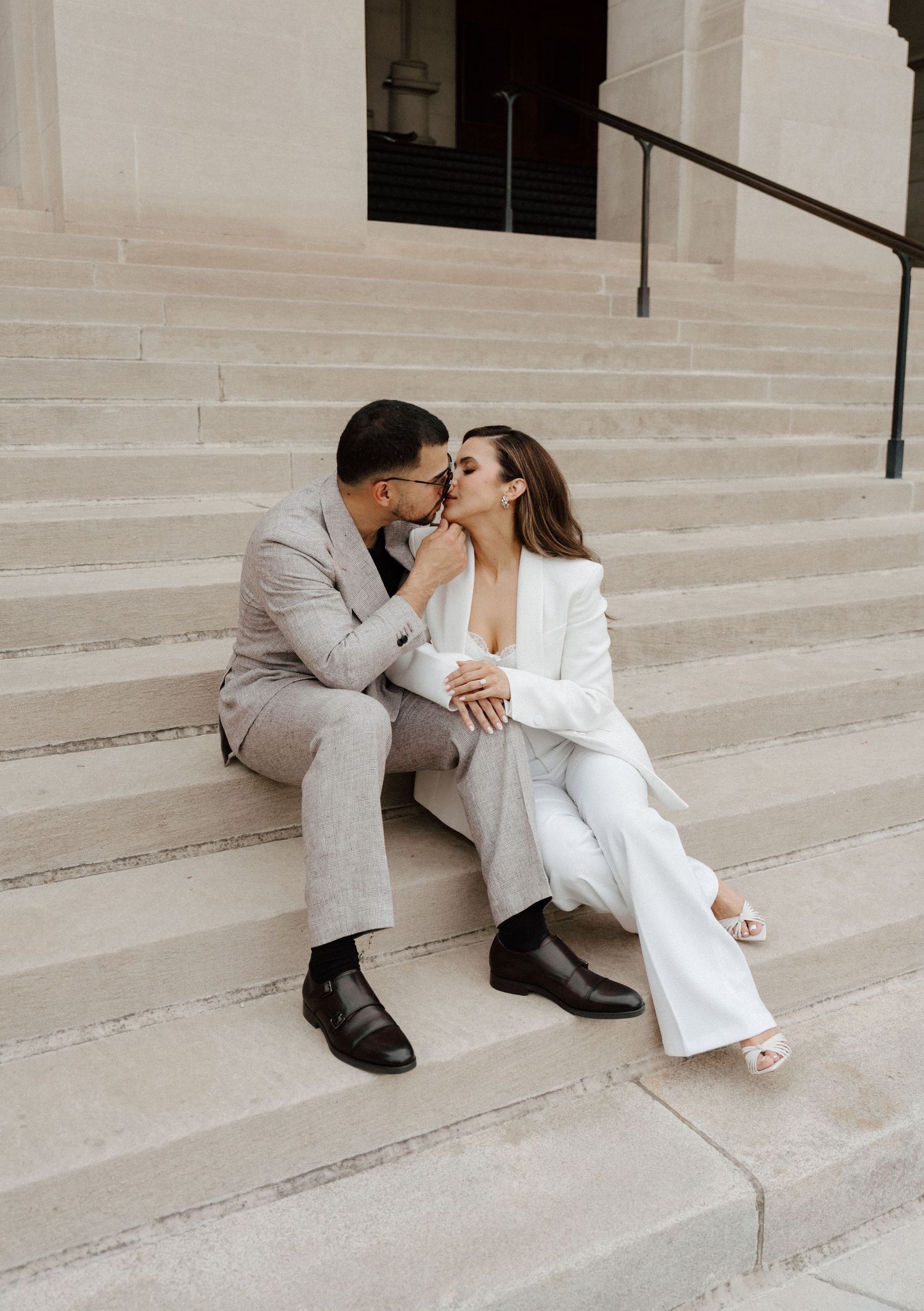 Couple sharing a kiss on stone steps, wearing elegant white suit and gray suit, in a romantic urban setting.