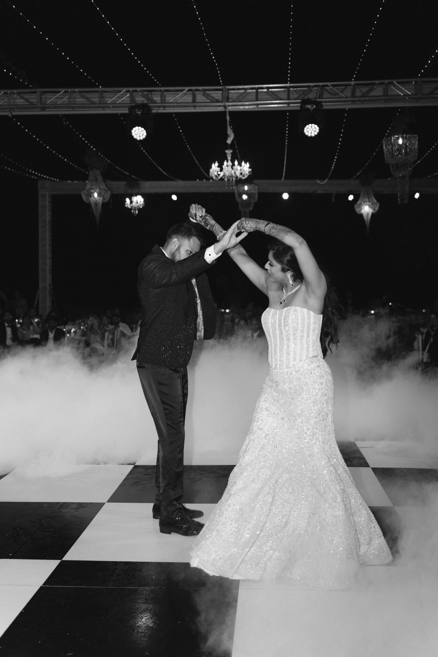 Bride and groom share first dance on checkered floor surrounded by fog under chandeliers in elegant black and white wedding photo.