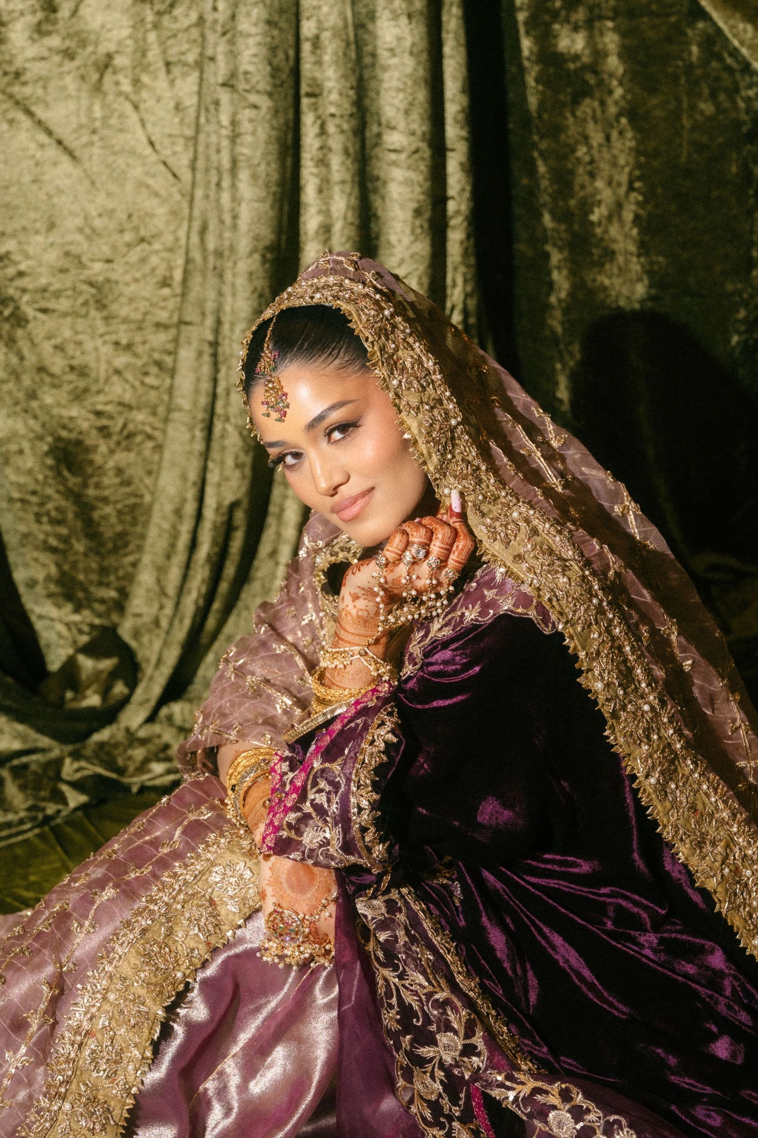 Indian bride in purple and gold lehenga with embroidered dupatta and traditional jewelry poses gracefully.