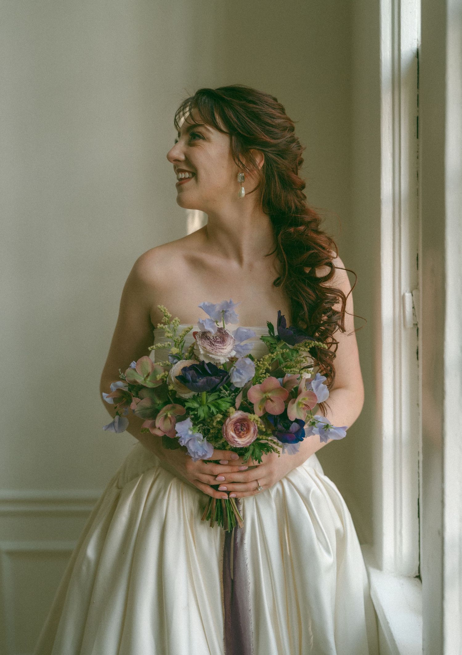 Smiling bride in strapless white gown holding colorful blue and pink floral bouquet beside a window.