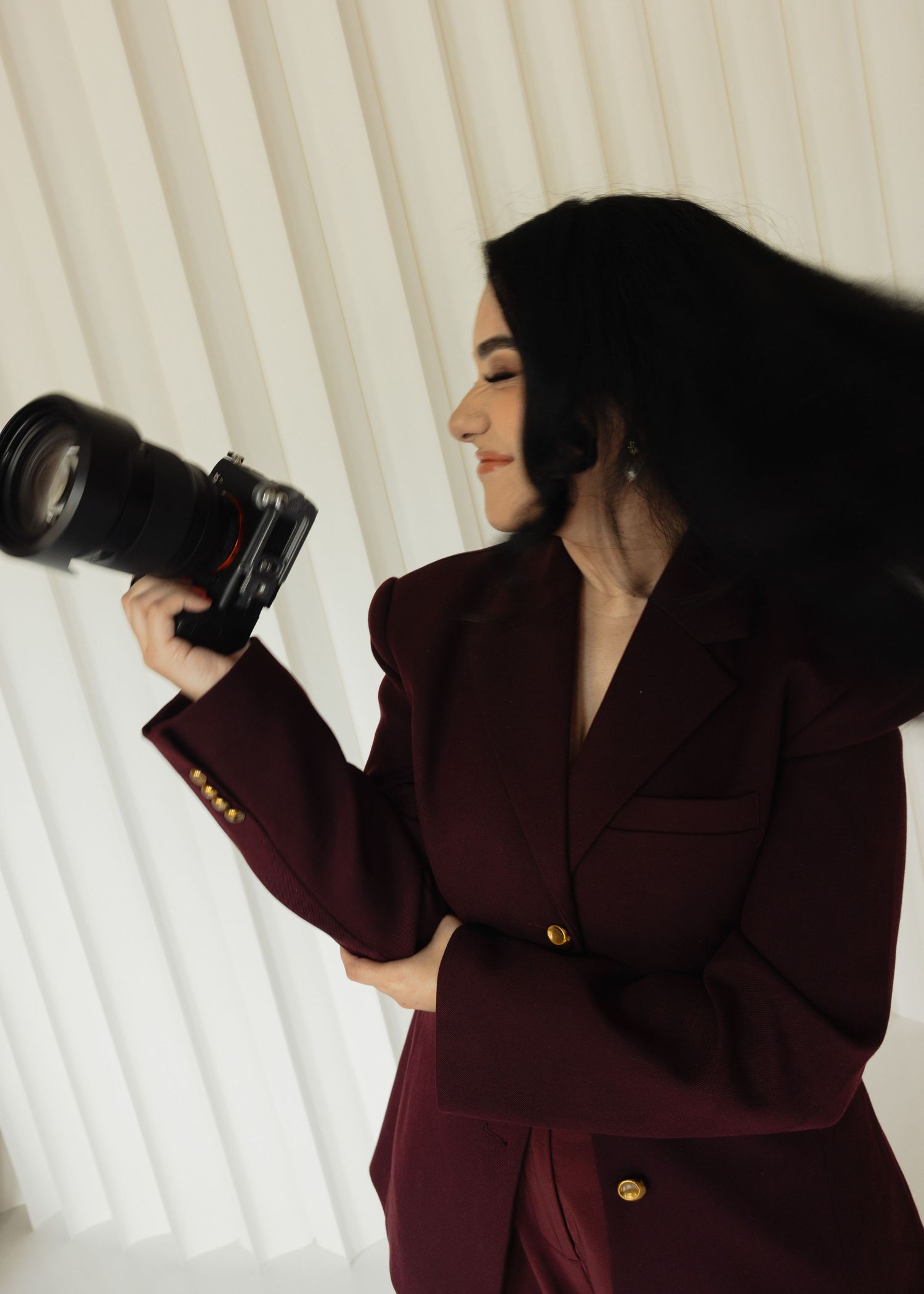 Woman in burgundy blazer holding a professional camera, posing against white curtains backdrop.