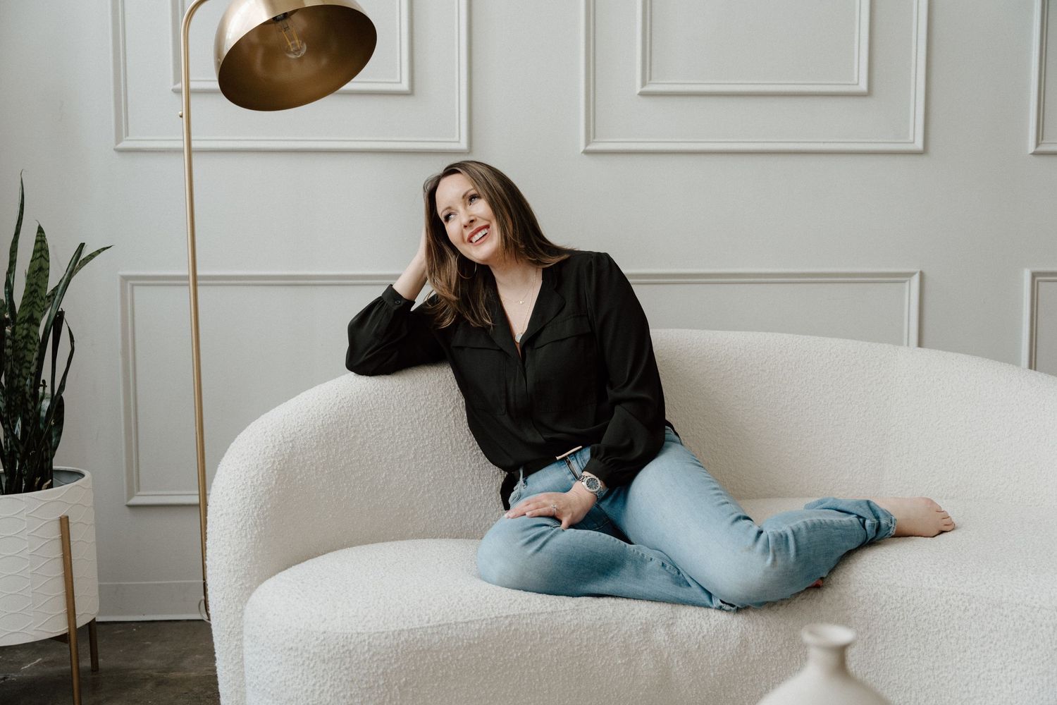 Woman in black blouse and jeans sitting relaxed on white sofa beside gold floor lamp and green plant.