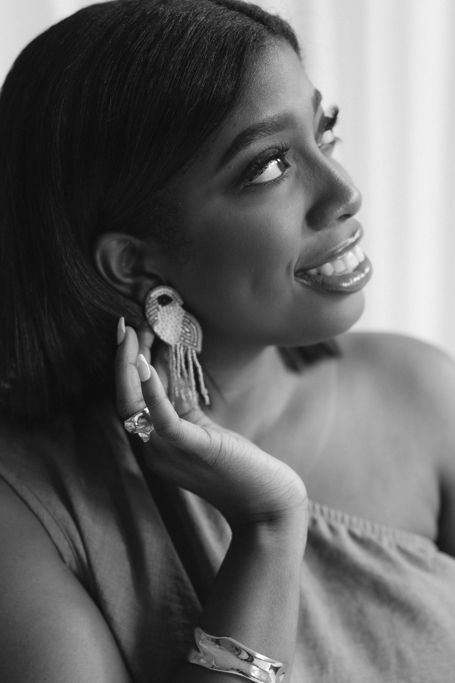 Black and white portrait of a smiling woman wearing elegant chandelier earrings and a bracelet, looking away.