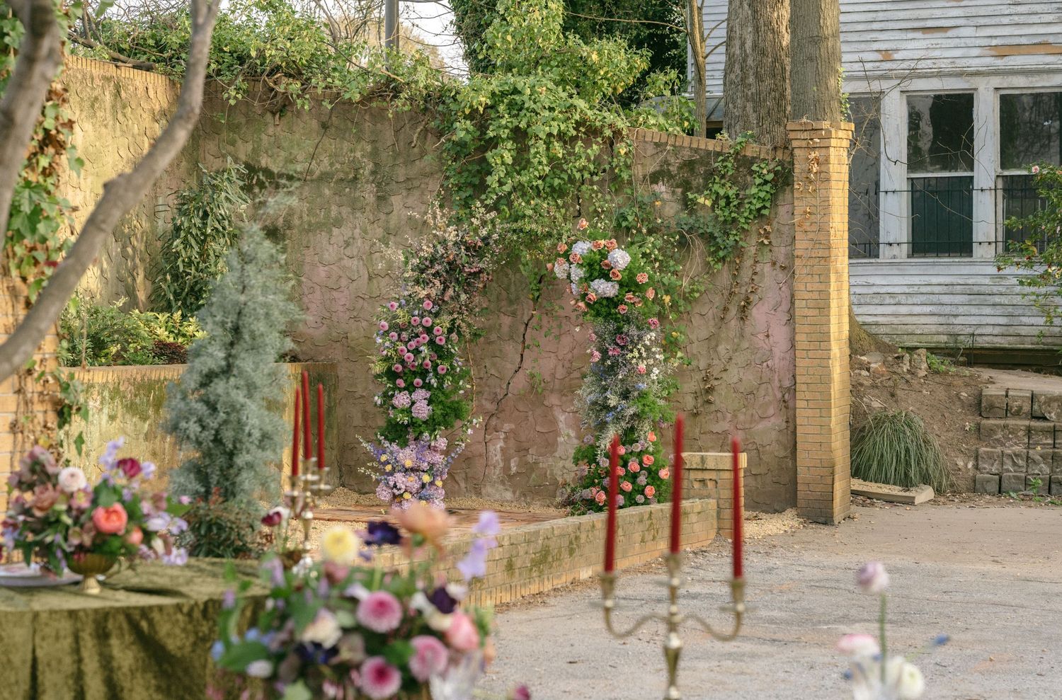 Romantic outdoor wedding setup with floral arch, pink roses, and rustic stone wall garden backdrop.
