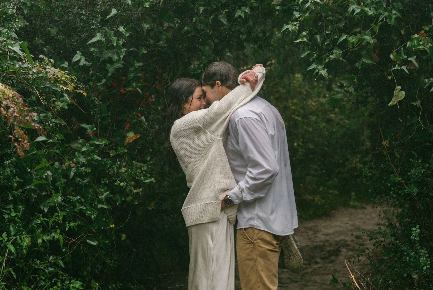 Jekyll Island Engagement. Couple embracing romantically in a lush green garden pathway, woman in white dress and man in light blue shirt.