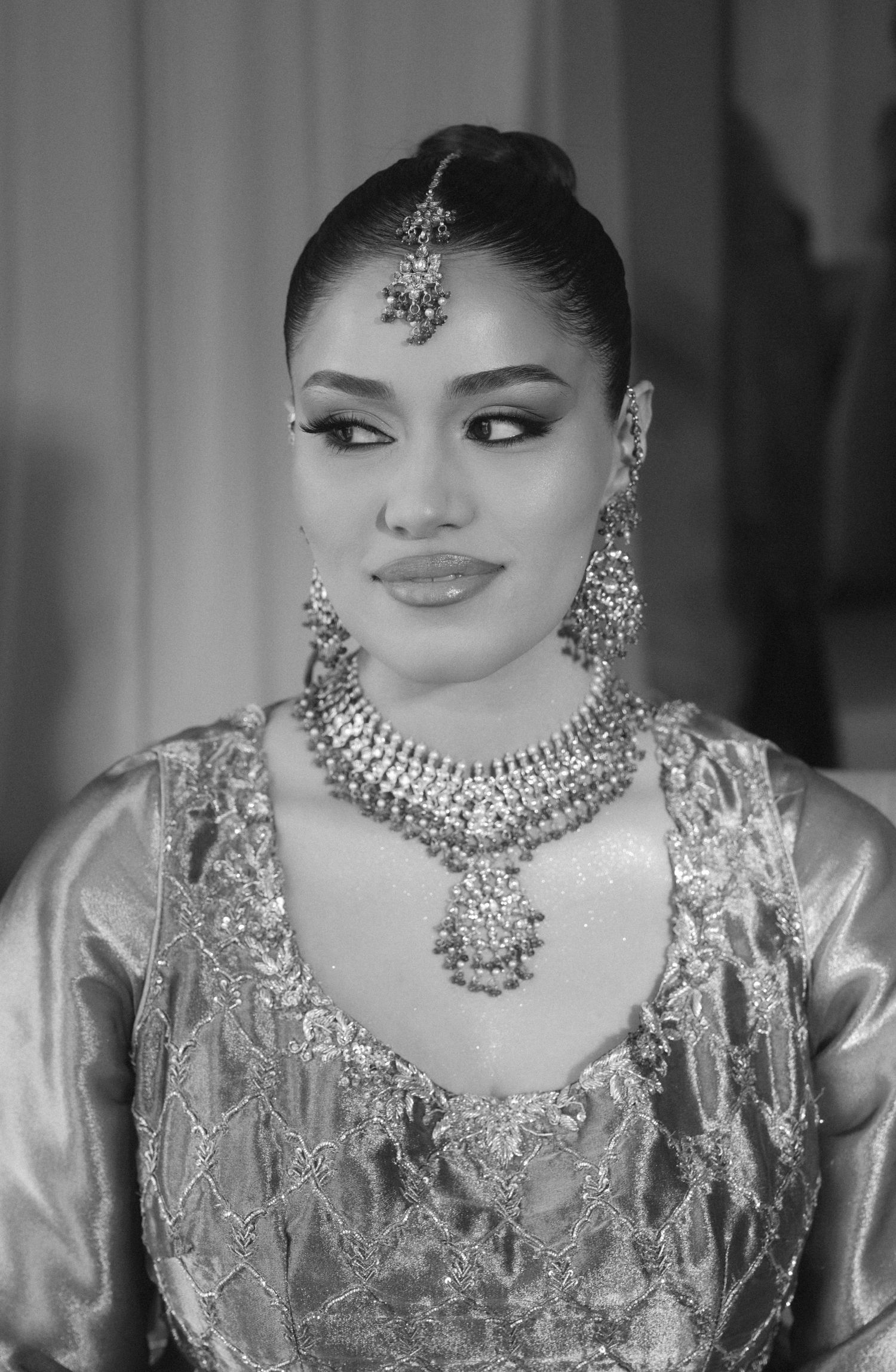Woman in traditional South Asian bridal jewelry, including maang tikka, jhumkas, and layered necklace, wearing embroidered attire.