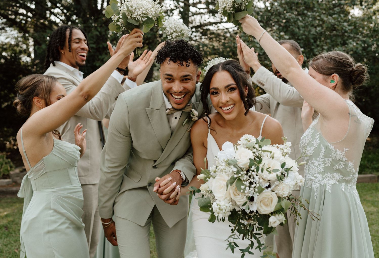 Joyful wedding party celebrates bride and groom outdoors, bridesmaids in sage green dresses, groom in tan suit.
