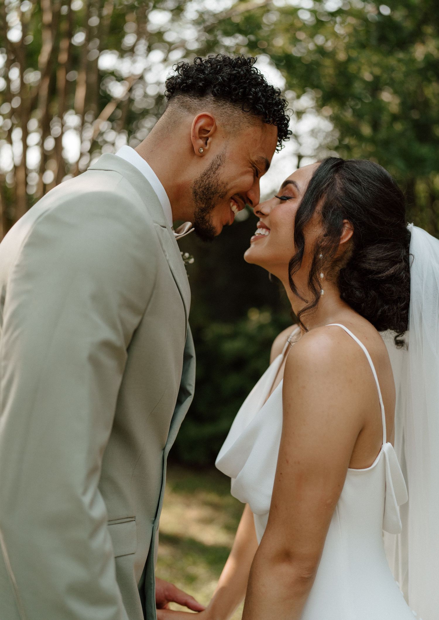 A joyful bride and groom in wedding attire share an intimate forehead-to-forehead moment, smiling outdoors among lush green trees.