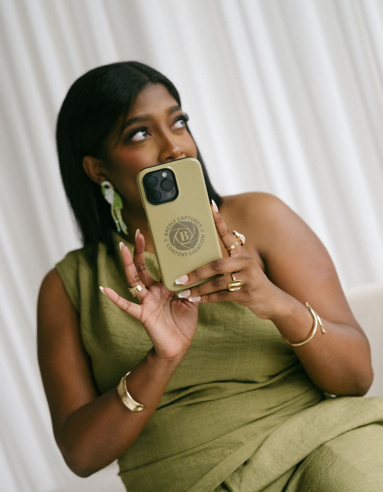 A Black woman in an olive green dress holds a gold phone case while looking away, wearing gold jewelry and green earrings.