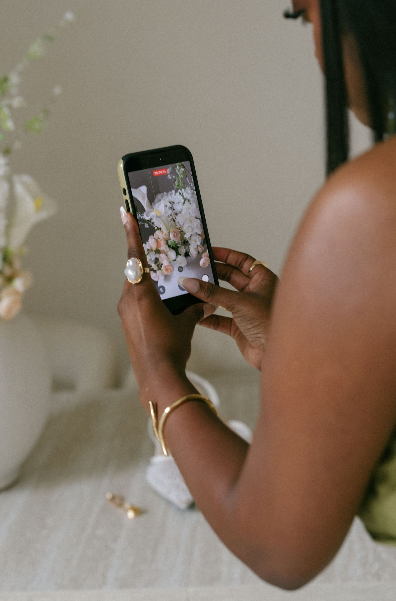 Woman photographing floral arrangement on smartphone wearing gold bracelet and pearl ring at event.