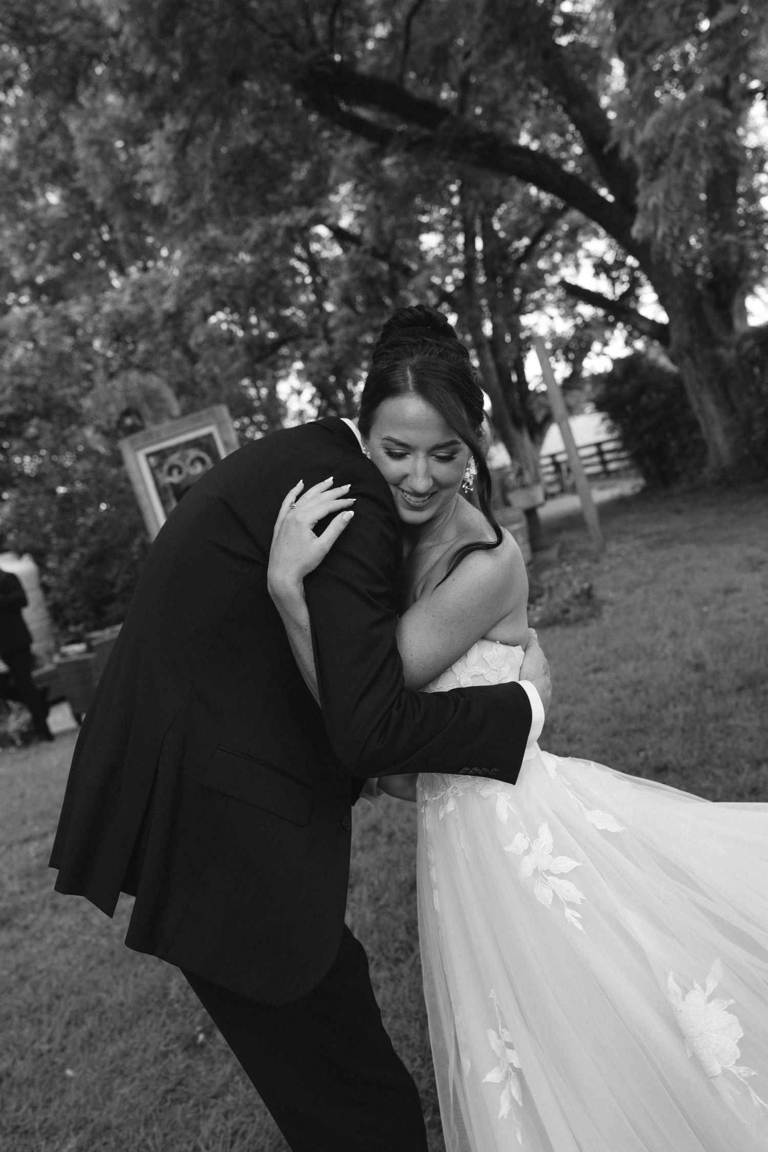 Black and white photo of a bride and groom sharing a joyful embrace outdoors at their wedding reception.