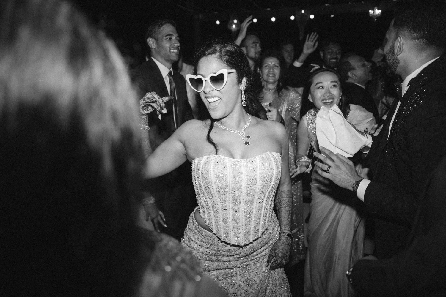 Woman in strapless gown and heart sunglasses dancing joyfully at a wedding reception in black and white photo.