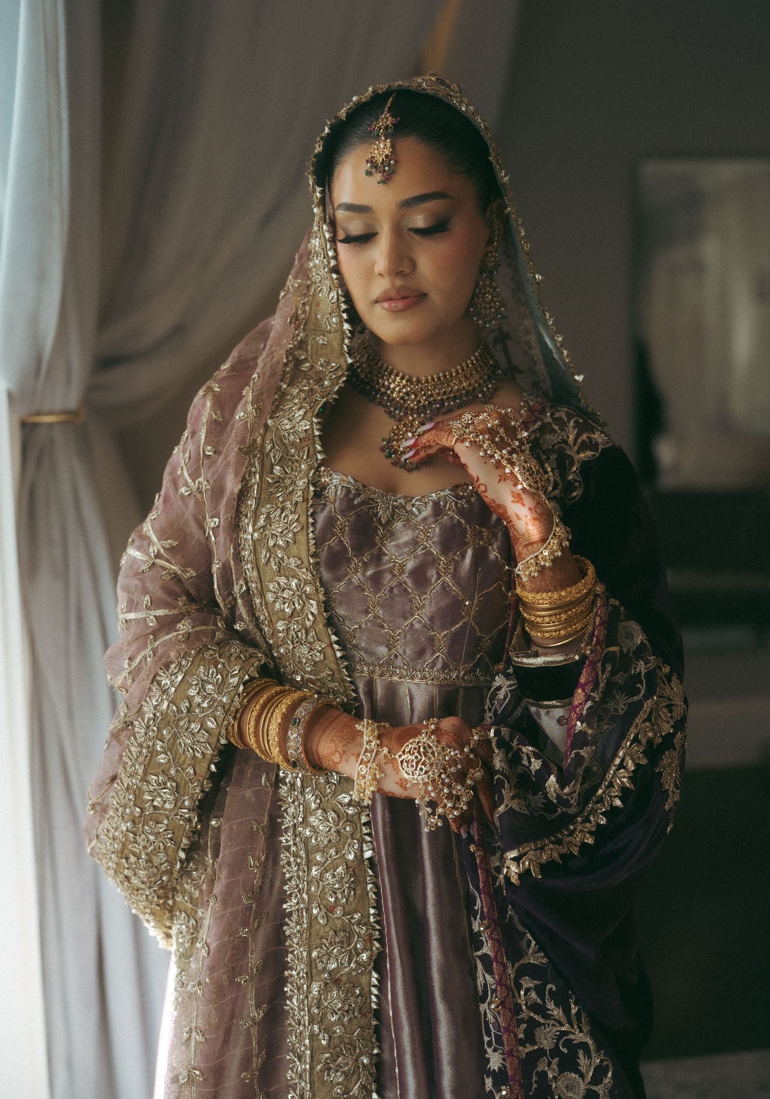 South Asian bride in mauve lehenga with gold embroidery, dupatta, bangles, and traditional jewelry poses elegantly.