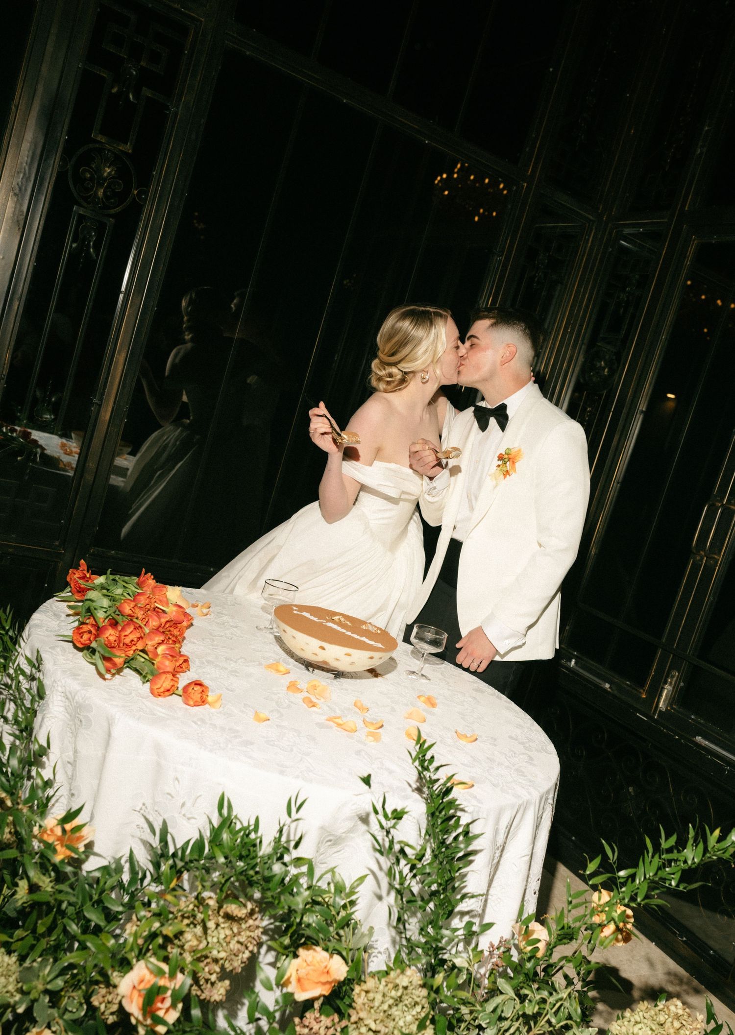 Bride and groom share a kiss at their wedding reception table adorned with orange florals and greenery at an elegant evening event.