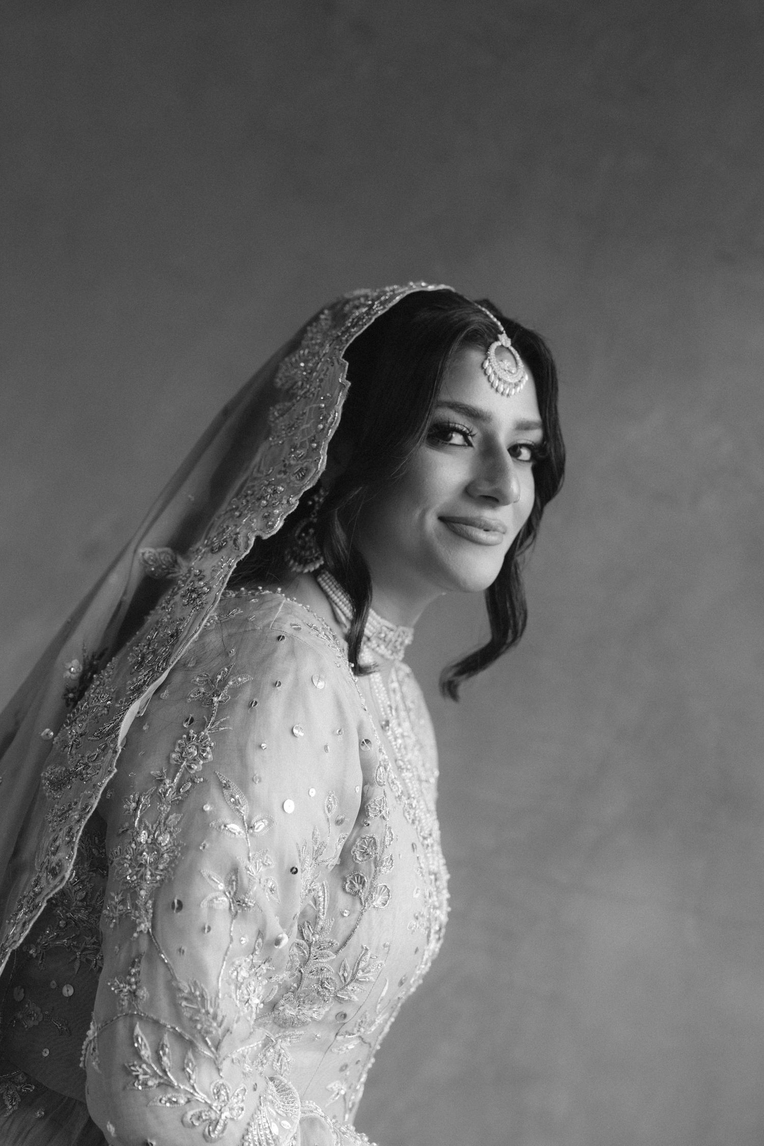 Black and white portrait of a smiling South Asian bride in embroidered bridal attire with veil and maang tikka jewelry.