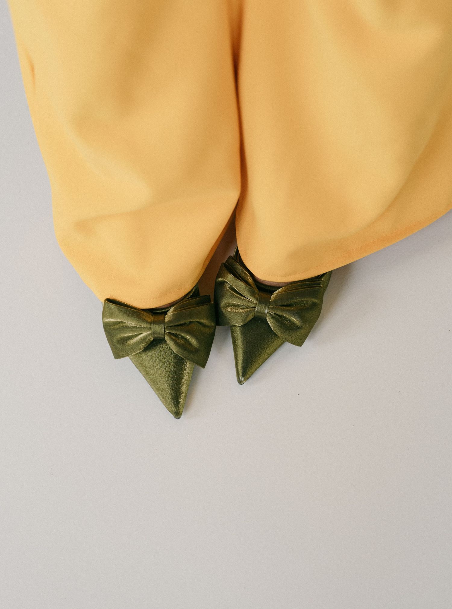 Green satin pointed-toe flats with bow detail peeking beneath yellow wide-leg trousers on grey background.