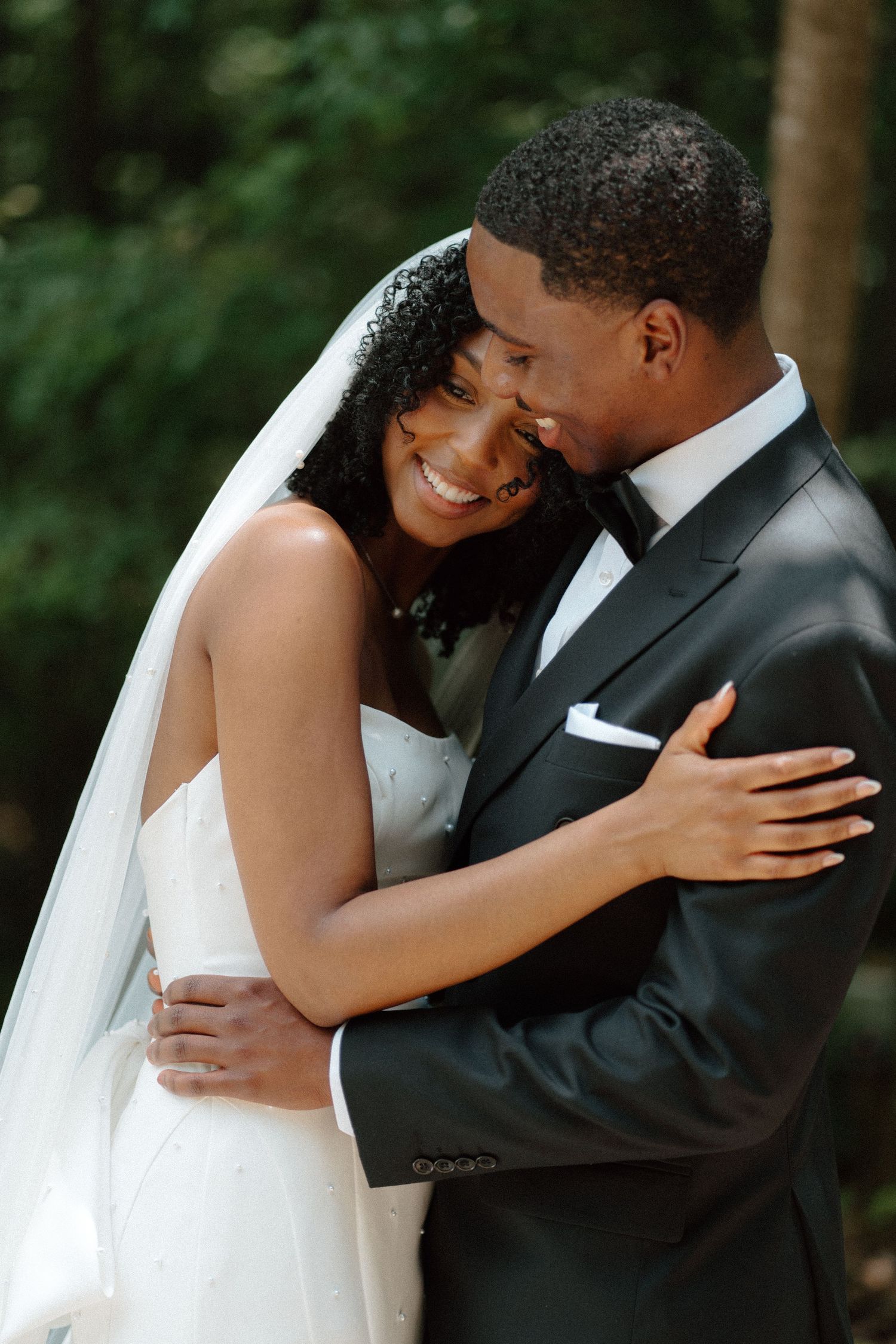 Happy bride in white gown and veil embracing groom in black tuxedo outdoors among trees.
