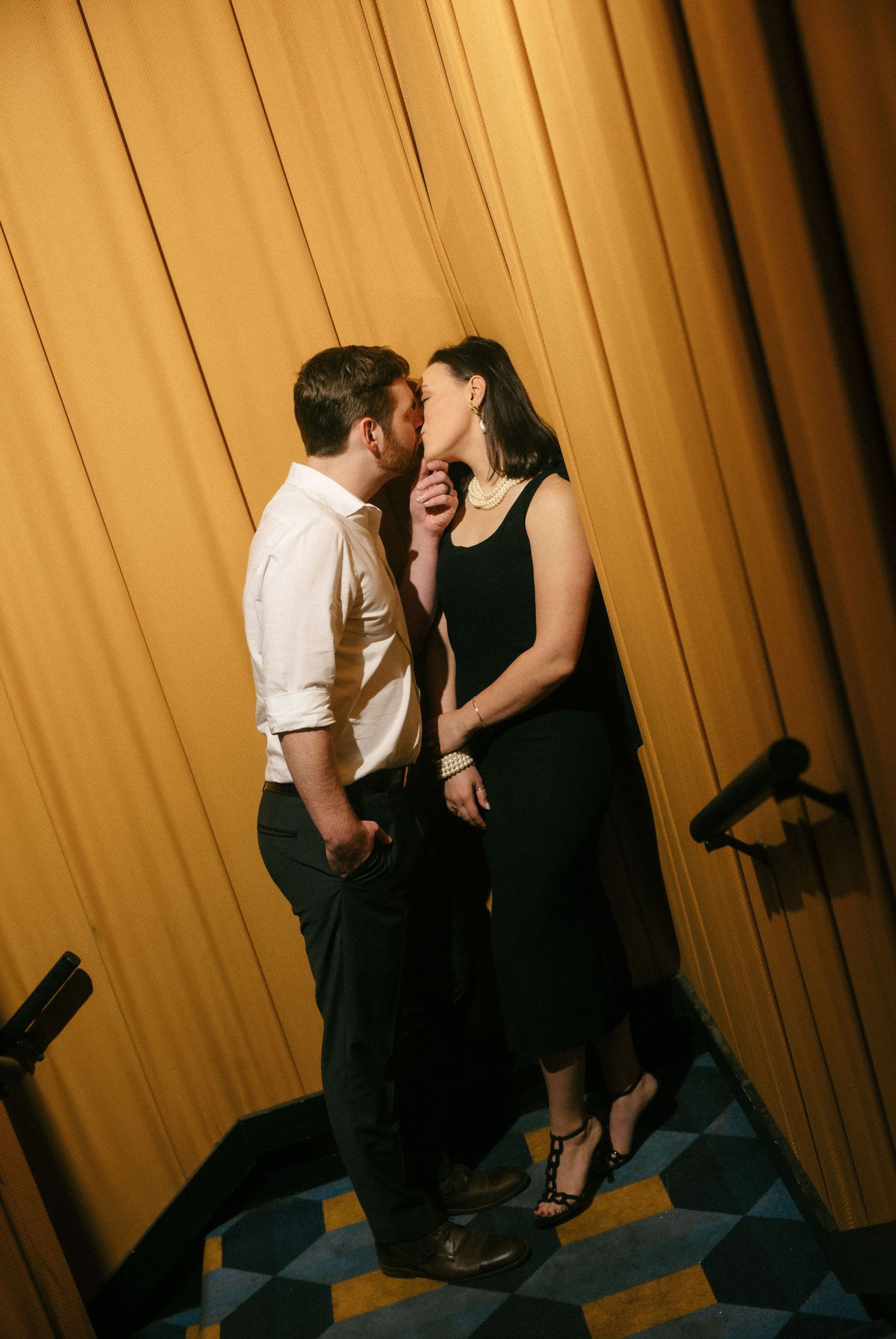 Couple sharing a romantic kiss in formal attire, man in white shirt and woman in black dress, standing by golden curtains. The Plaza Theatre